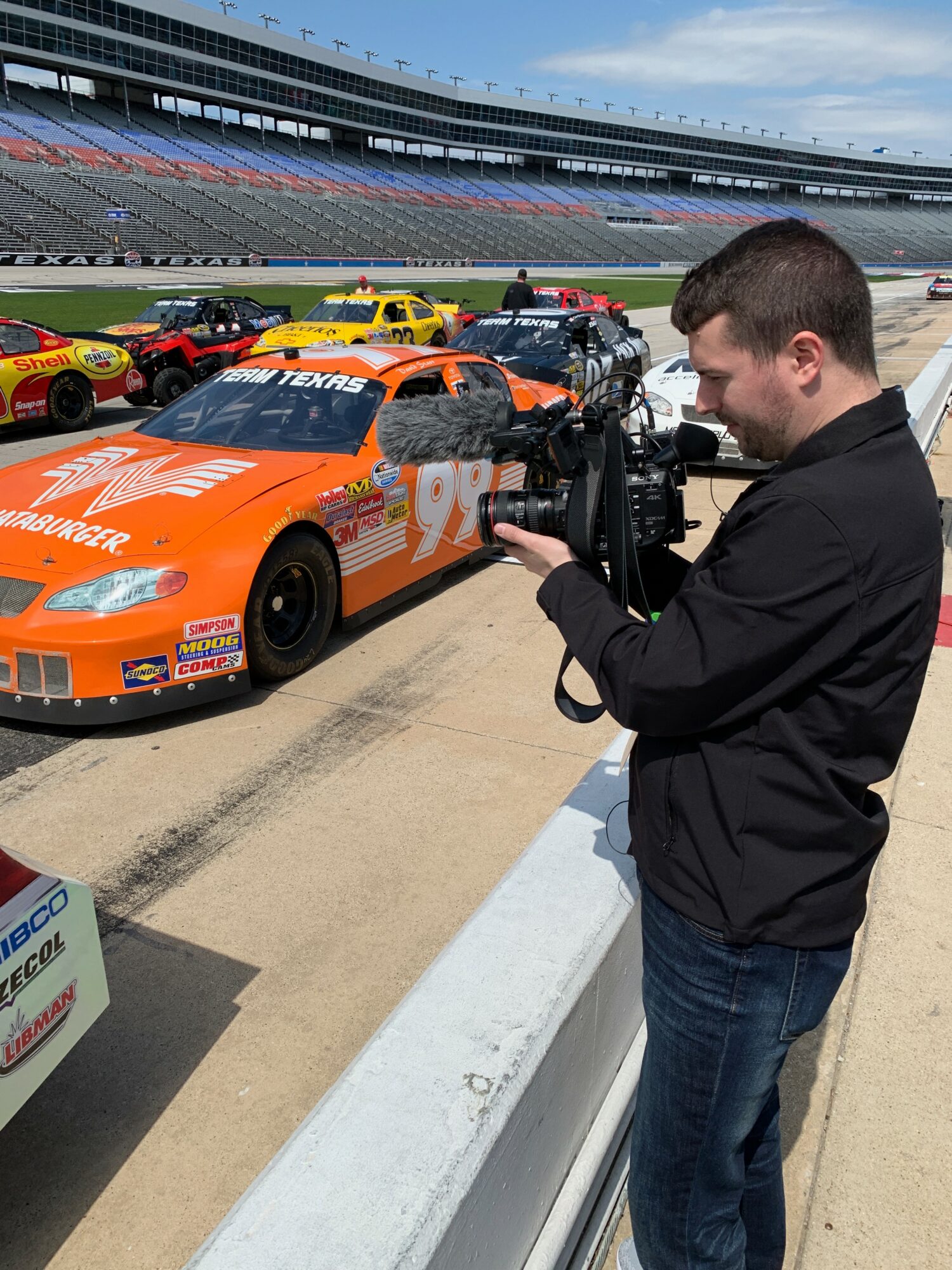 Person filming race cars on track with empty grandstands in background, sunny day, racing event setting.