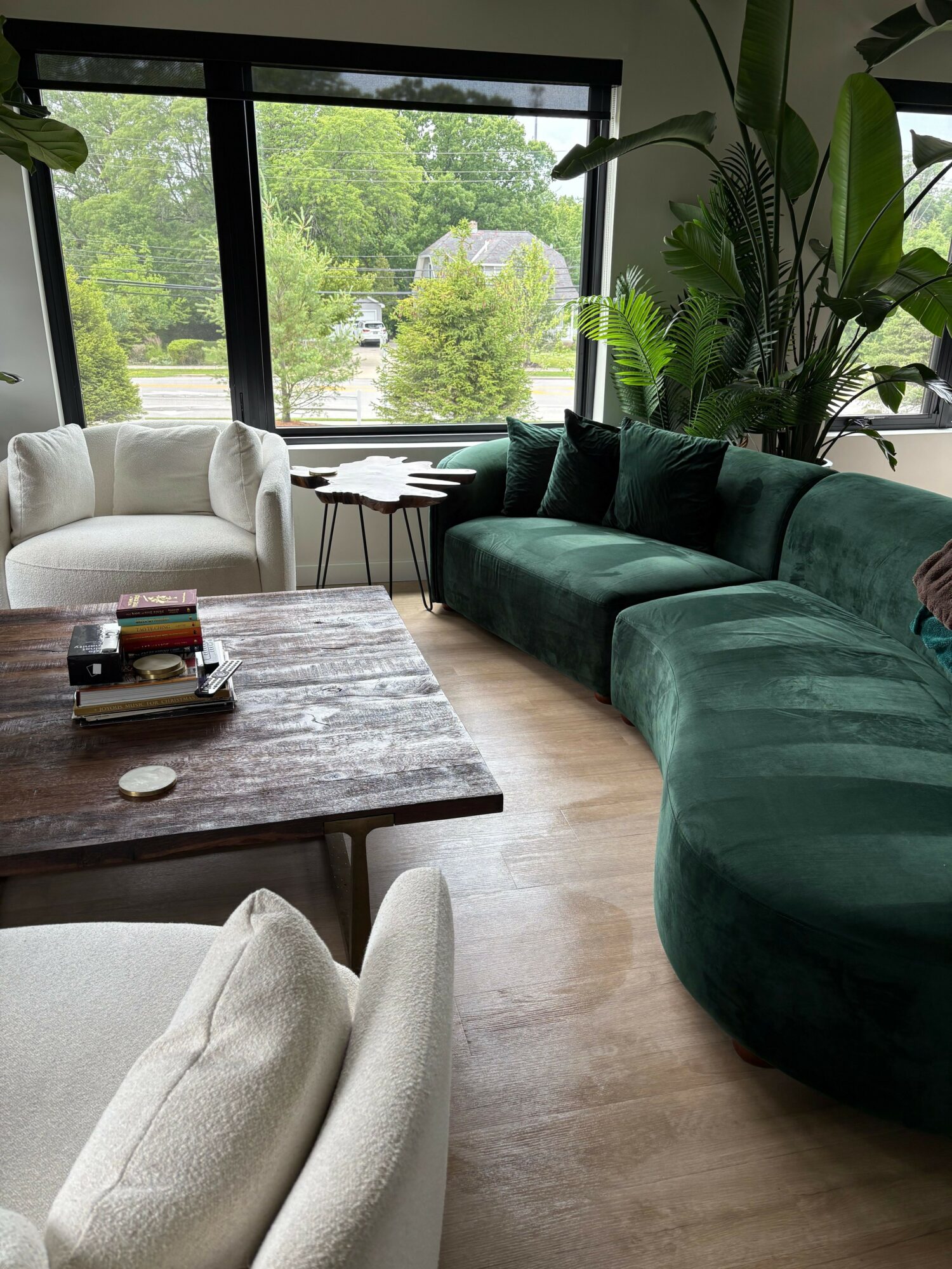 Living room with large window, green sofa, white armchair, wooden coffee table, and view of trees outside.