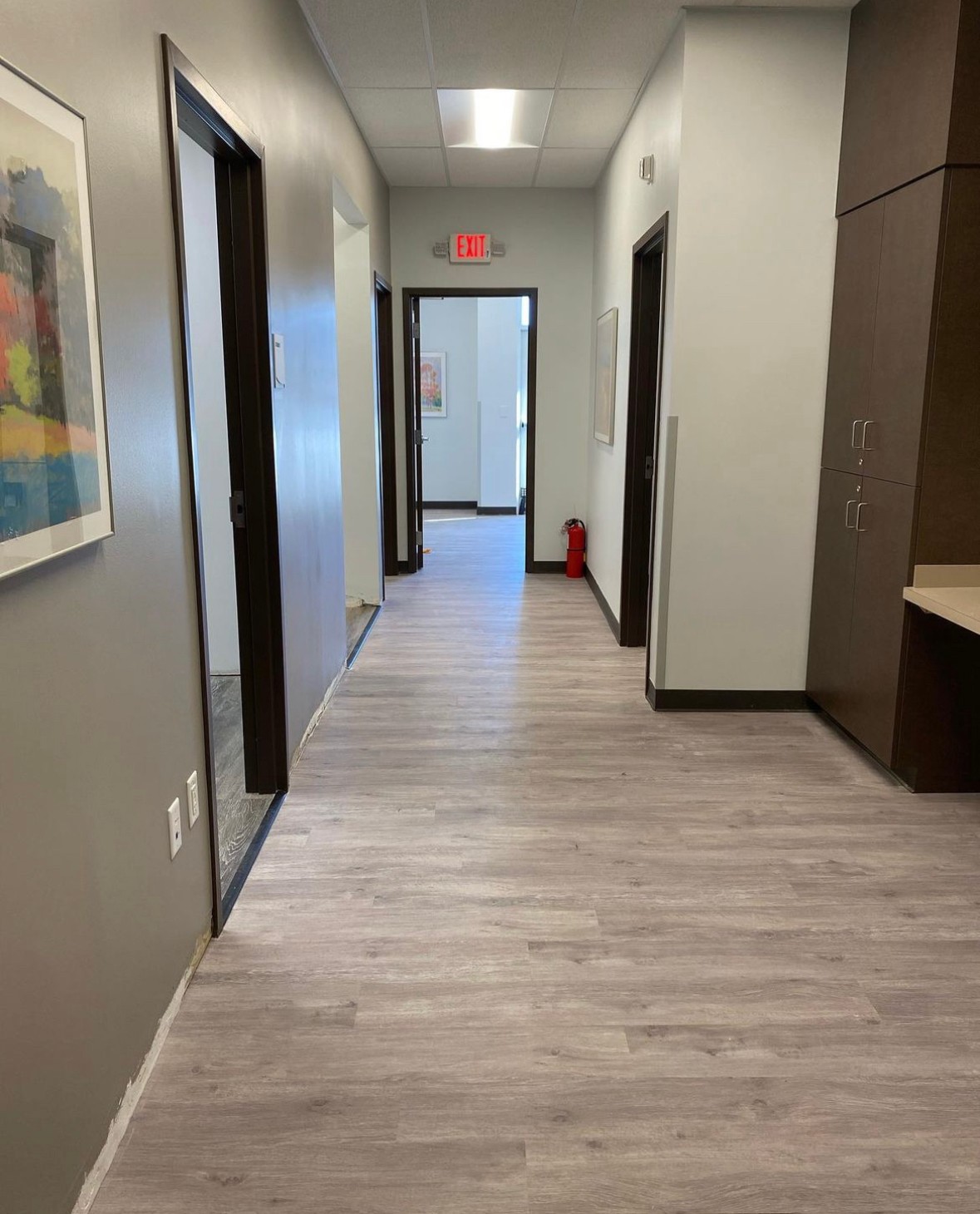 Hallway with light-colored wood flooring, white walls, and multiple doors, leading to a room with a fire extinguisher visible.