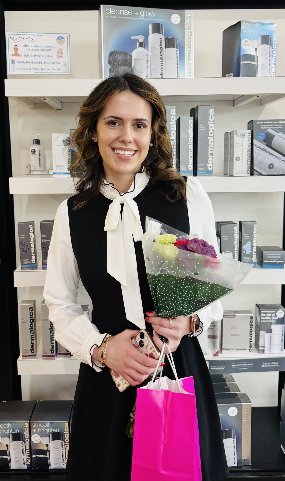 Woman with brown hair holding a bouquet of flowers and a pink shopping bag, standing in front of a bookshelf.