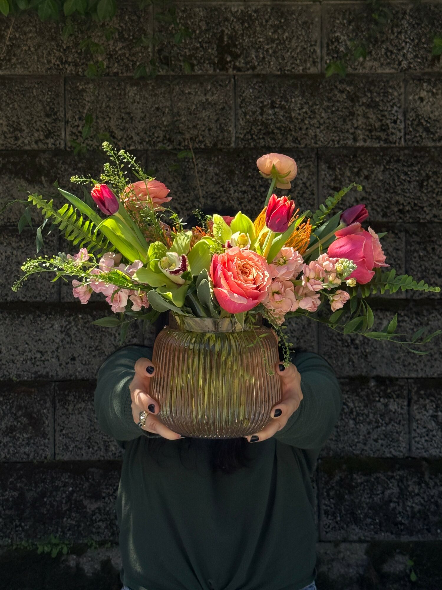 Person holding a large flower arrangement in a brown vase with pink, red, and purple flowers, against a dark brick wall.
