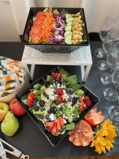 Two black square bowls with colorful salads and vegetables, surrounded by fruits, flowers, and glasses on a black table.