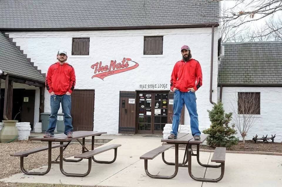 Two men in red jackets stand on picnic tables outside a building with a sign that reads 'The Mel's'.