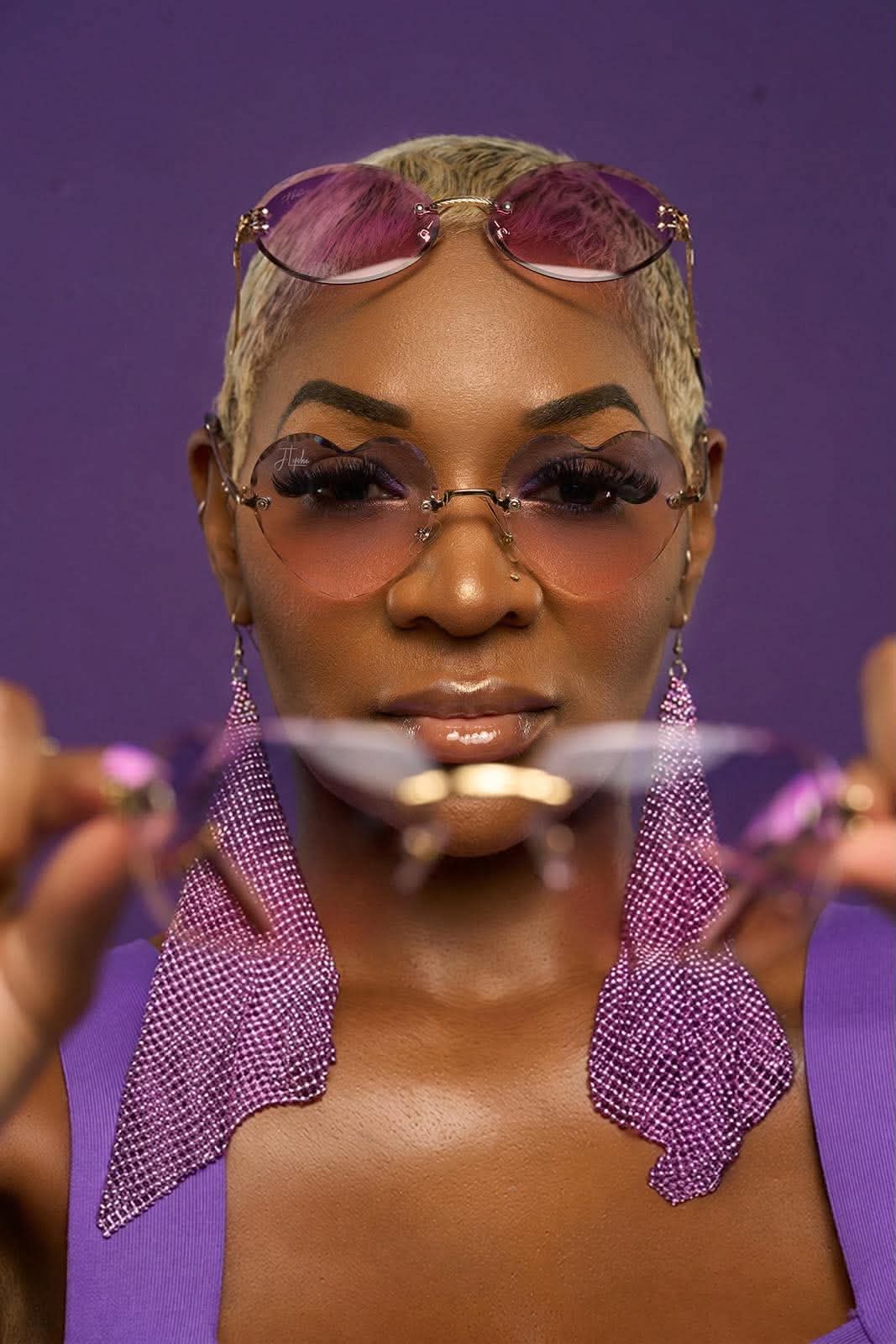 Woman with short blonde hair, sunglasses on head, holding a spoon close to face, purple background, wearing purple top.