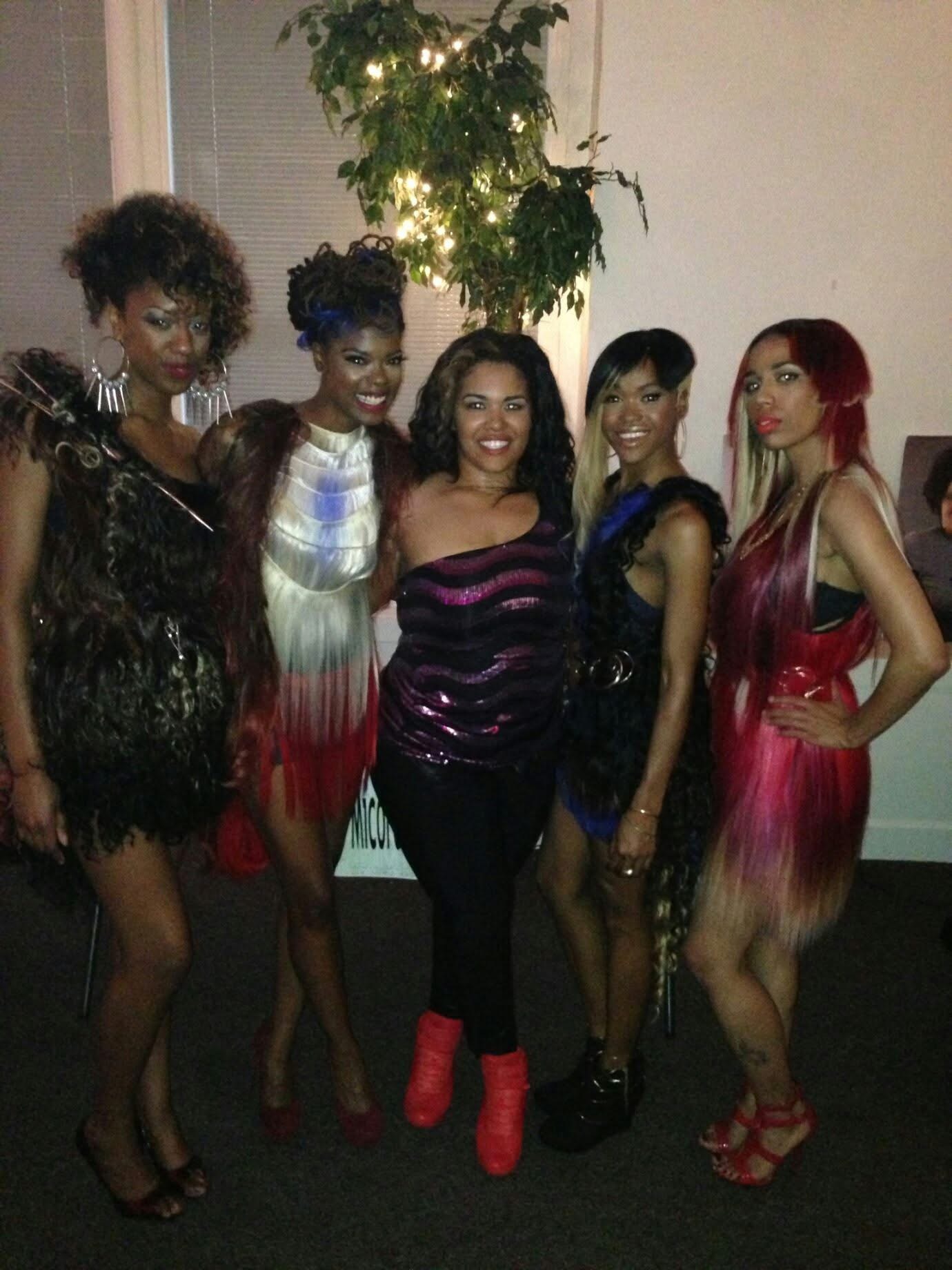 Five women in colorful dresses and heels pose indoors with a plant and light in the background.