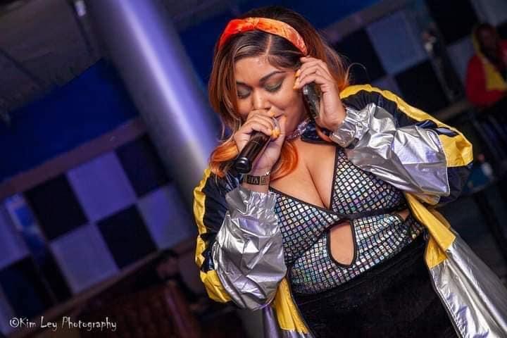 Young woman singing into a microphone, wearing a colorful jacket and patterned top, in a dimly lit setting.