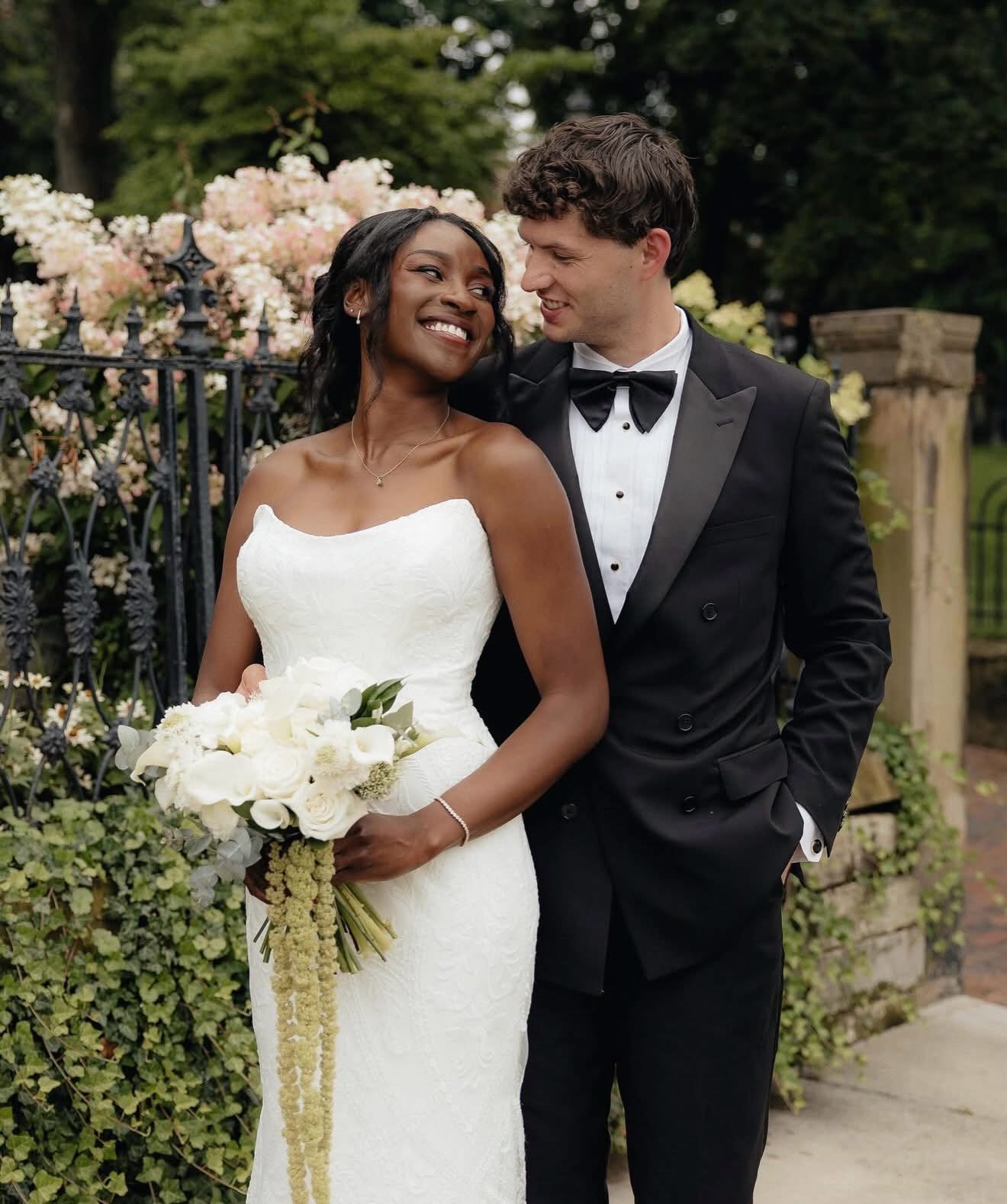 Couple in wedding attire outdoors, smiling, with greenery and flowers in background.