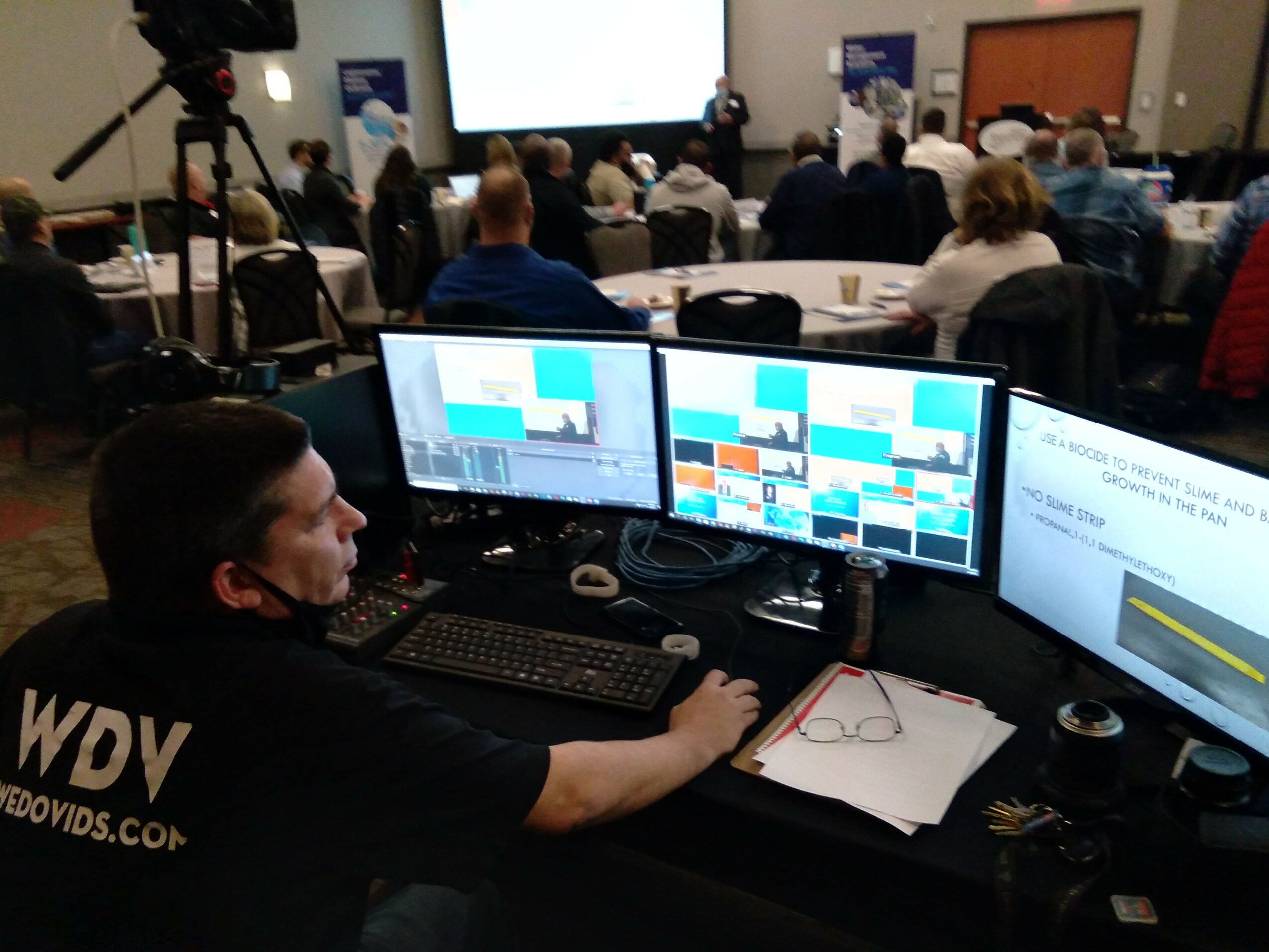 Conference room with attendees, presentation screens, and a person working at a desk with multiple monitors.