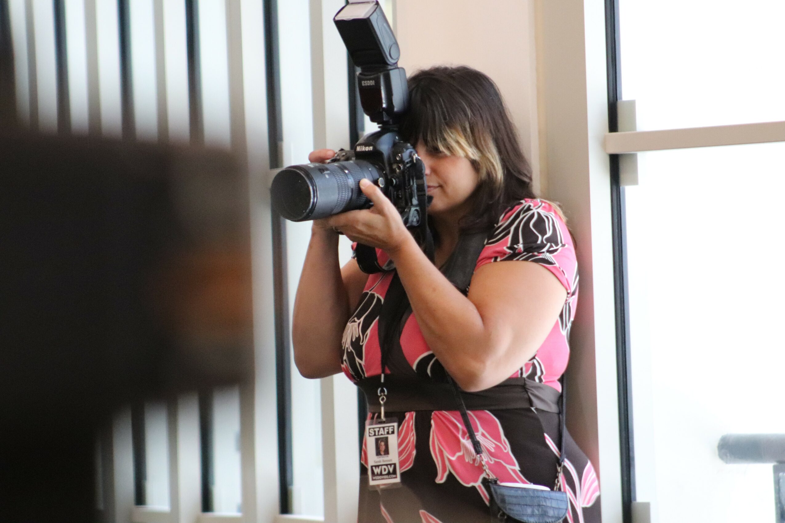 Woman taking a photo with a camera and flash, standing indoors near a window, wearing a pink and black patterned dress.