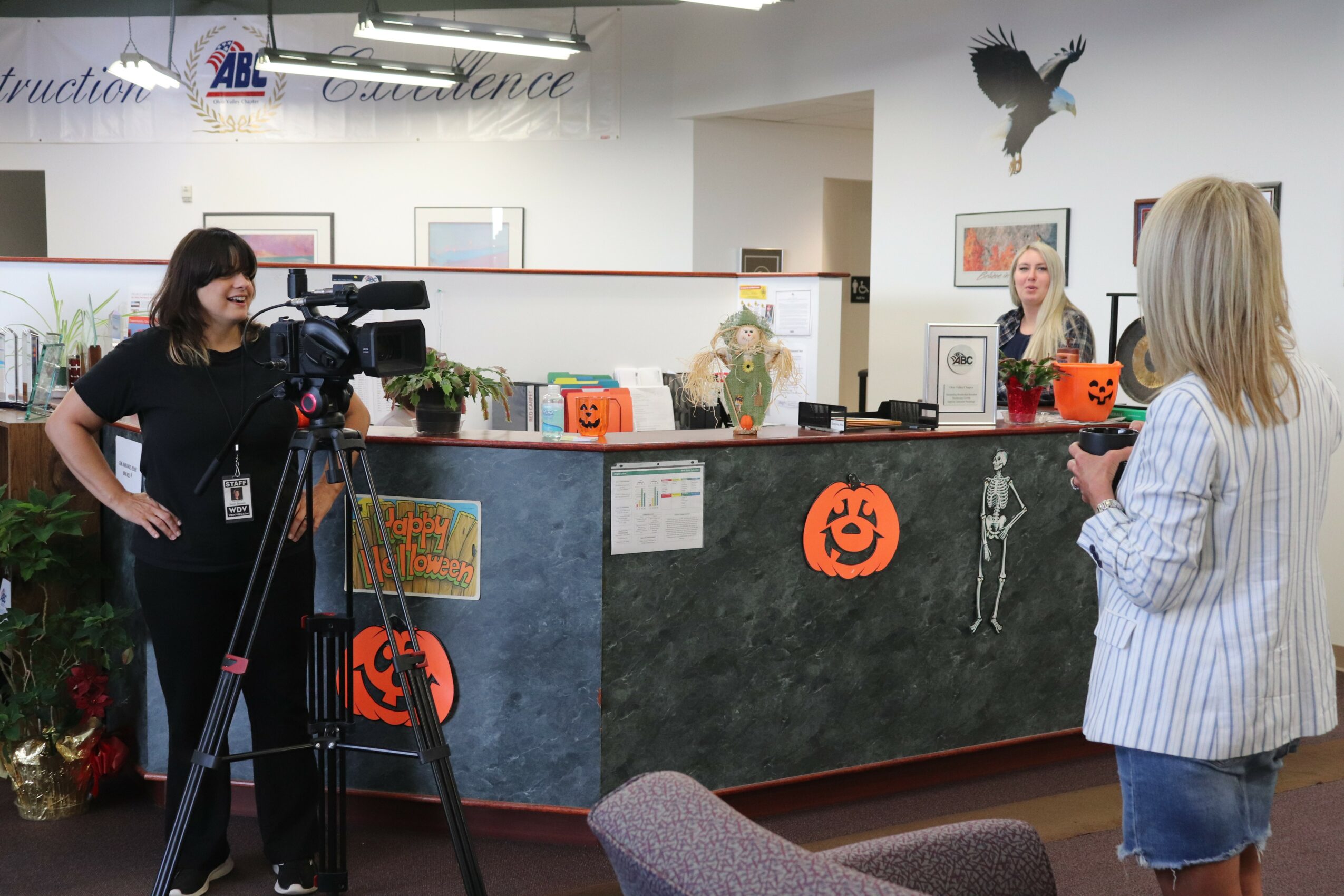 Two women at a reception desk with Halloween decorations, including pumpkins and a skeleton, in an office setting.