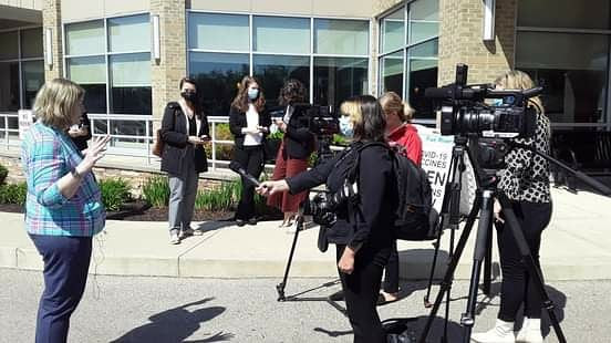 Group of people, some with cameras, gathered outside a building with large windows, sunlight, and shadows.