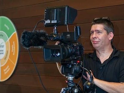 Man operating a professional video camera indoors, wooden wall background