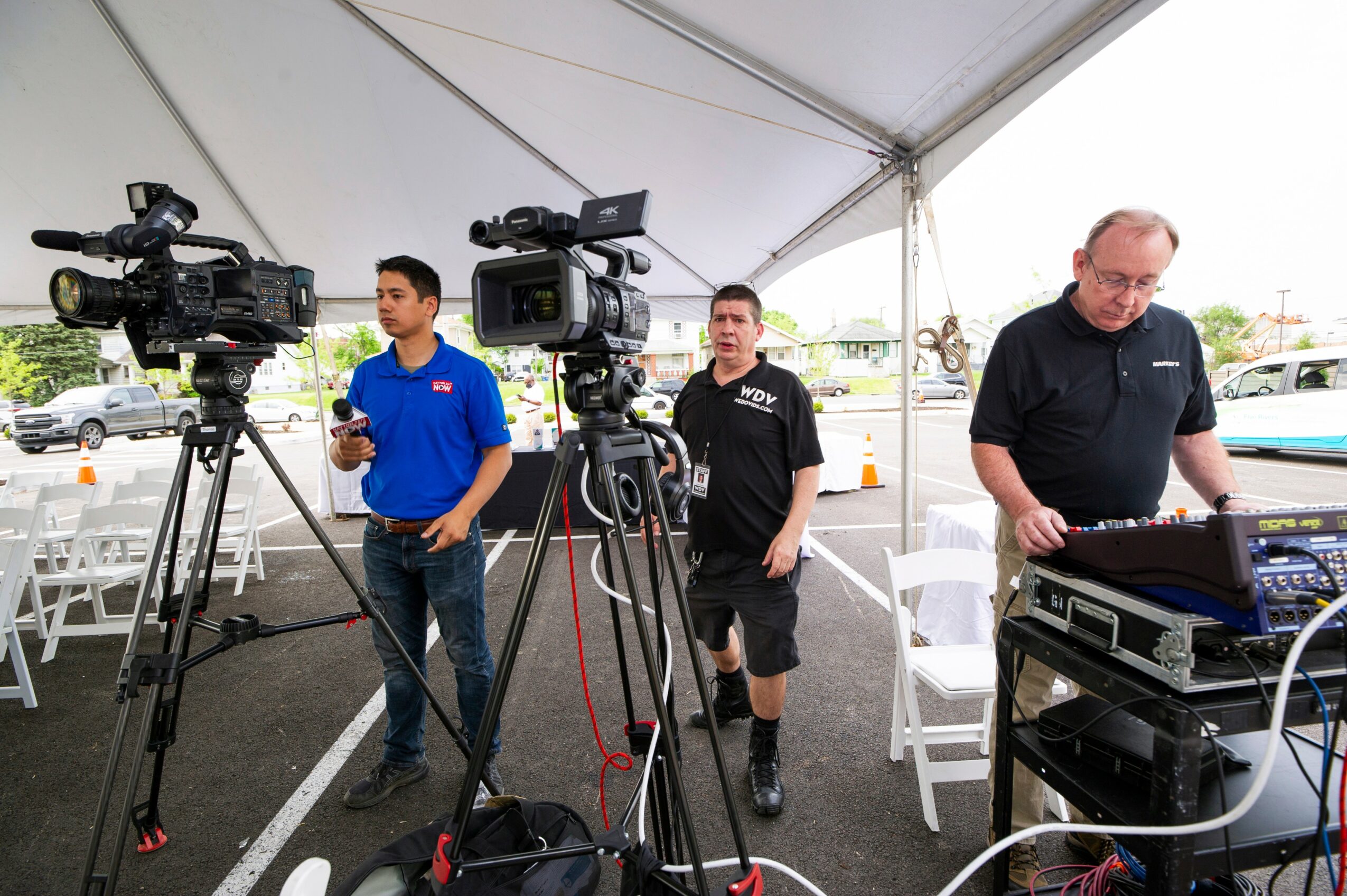 Three men under a tent with cameras and audio equipment, preparing for a recording or broadcast, with chairs and a parking lot in background.