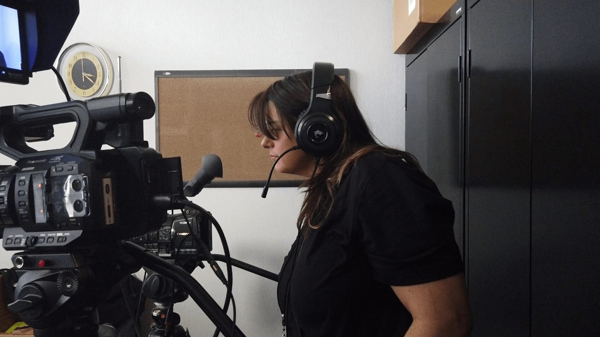 Woman wearing headphones sits in front of camera equipment in a room with black cabinets and a corkboard.