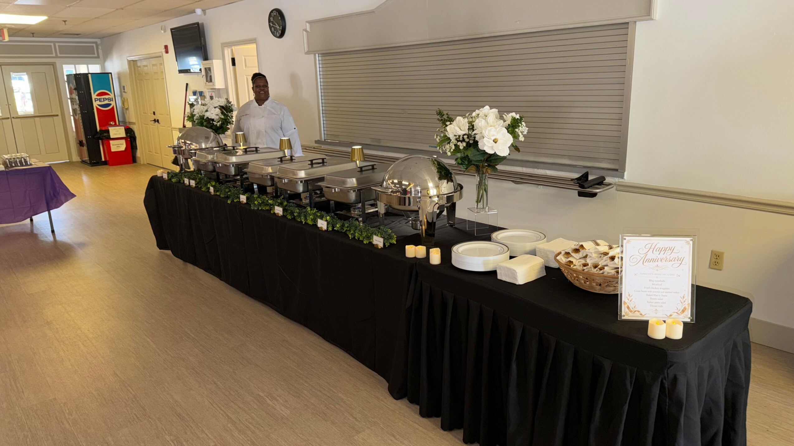 Buffet table with food, flowers, and a person behind it in a room with light-colored walls and wooden floor.