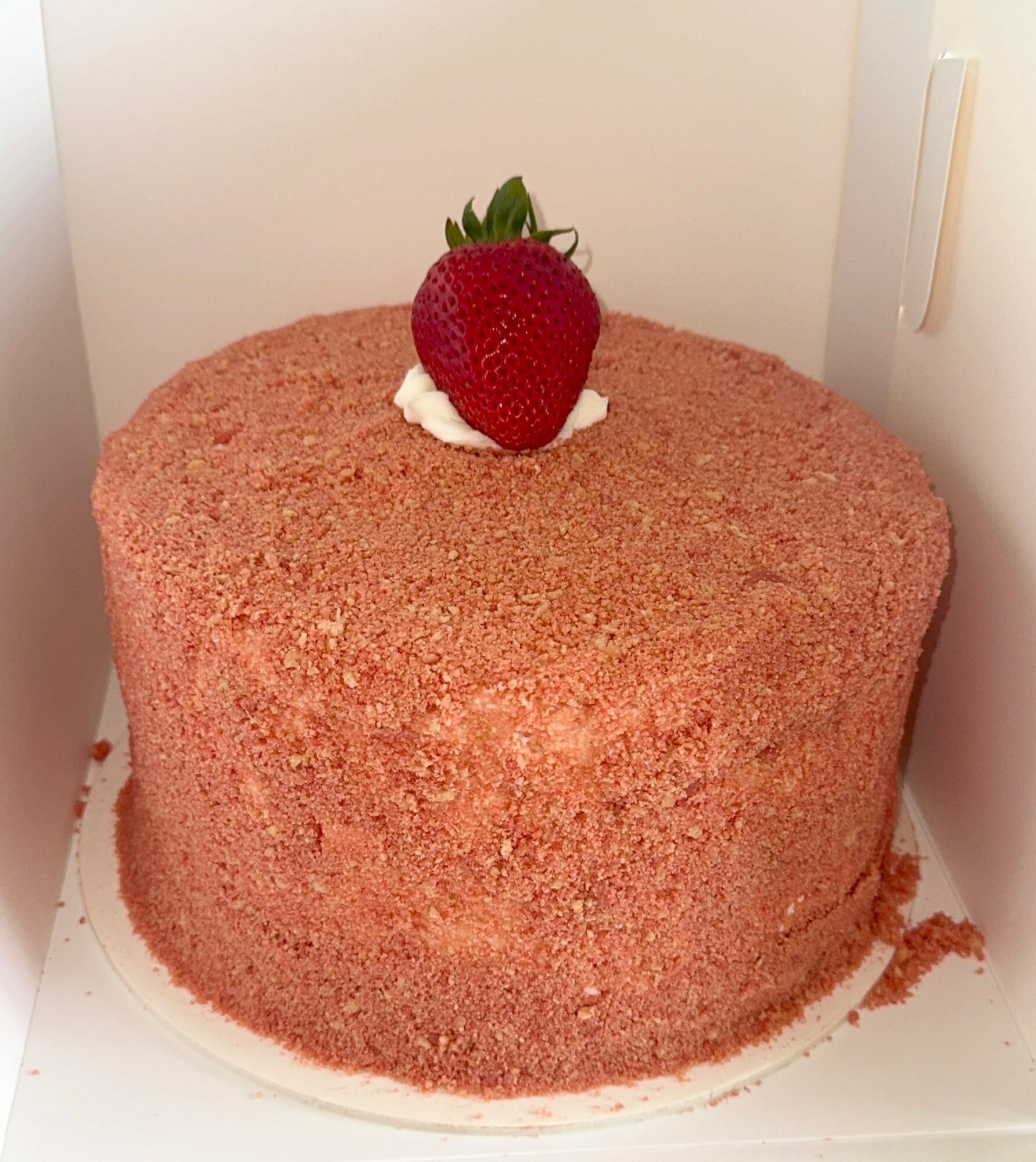 Round pink cake with a strawberry on top, inside a white box, with crumbs around the base.