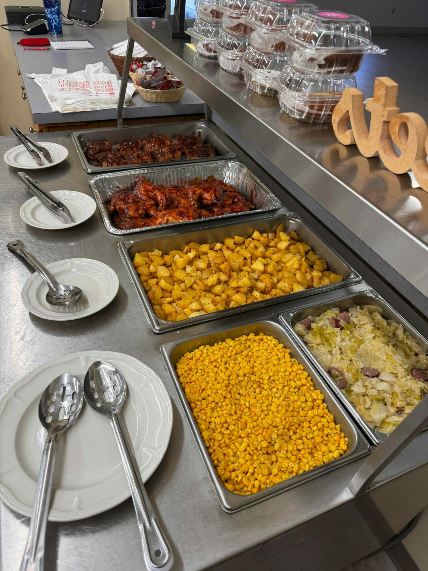 Buffet with three trays of food: corn, mashed potatoes, and a mixed dish, with plates and serving utensils nearby.