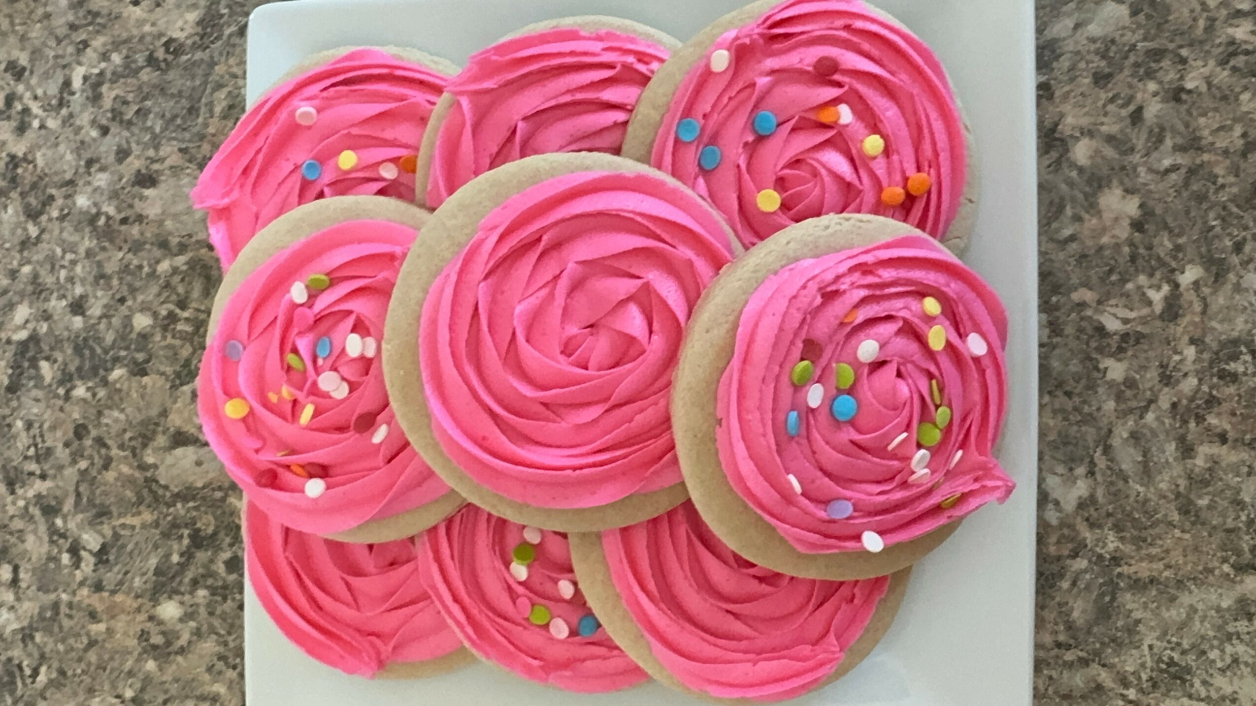 Plate of seven cupcakes with pink frosting and colorful sprinkles, on a gray surface.