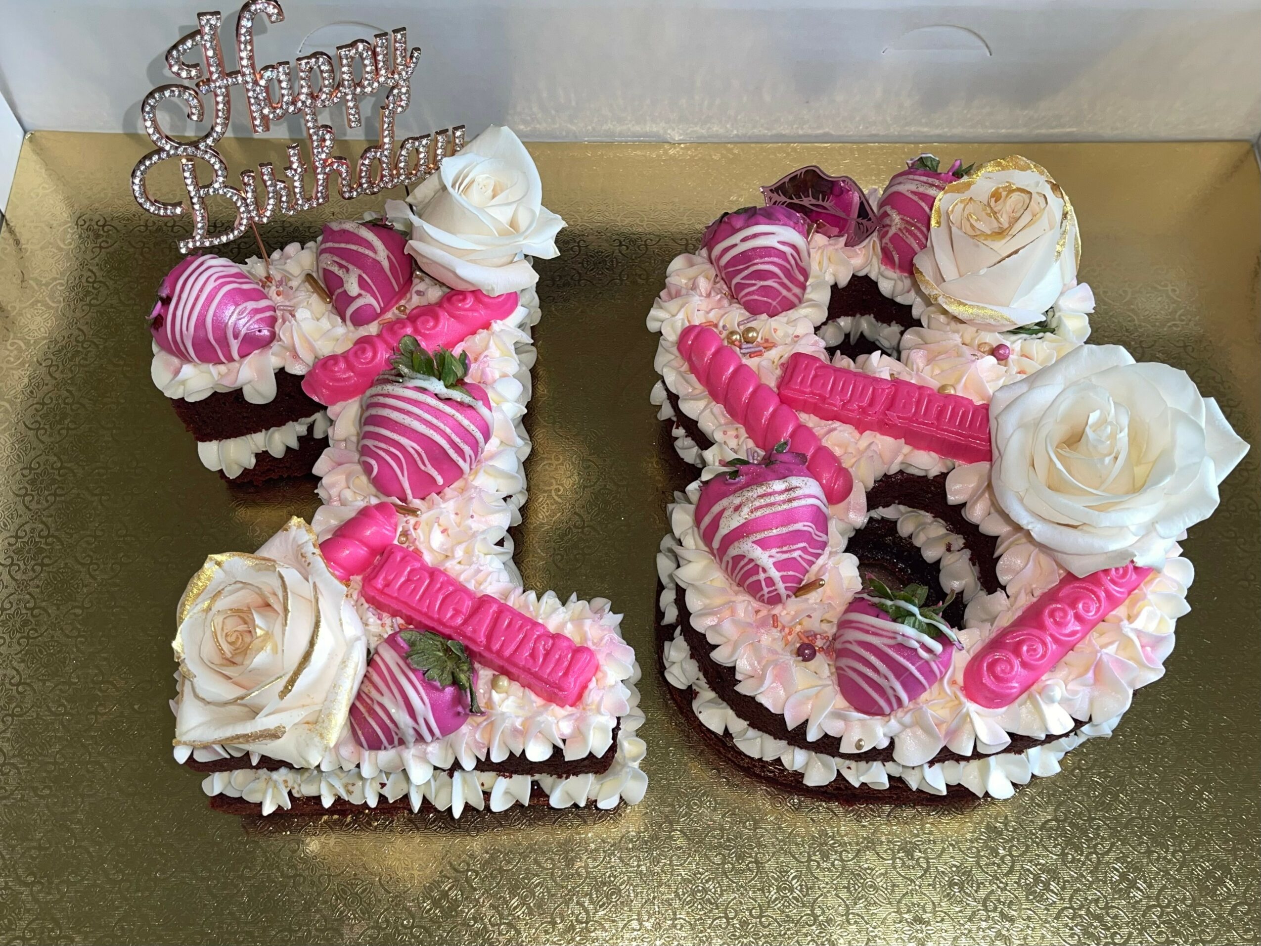 Number-shaped birthday cake decorated with pink and white flowers, hearts, and a 'Happy Birthday' topper, on a gold surface.