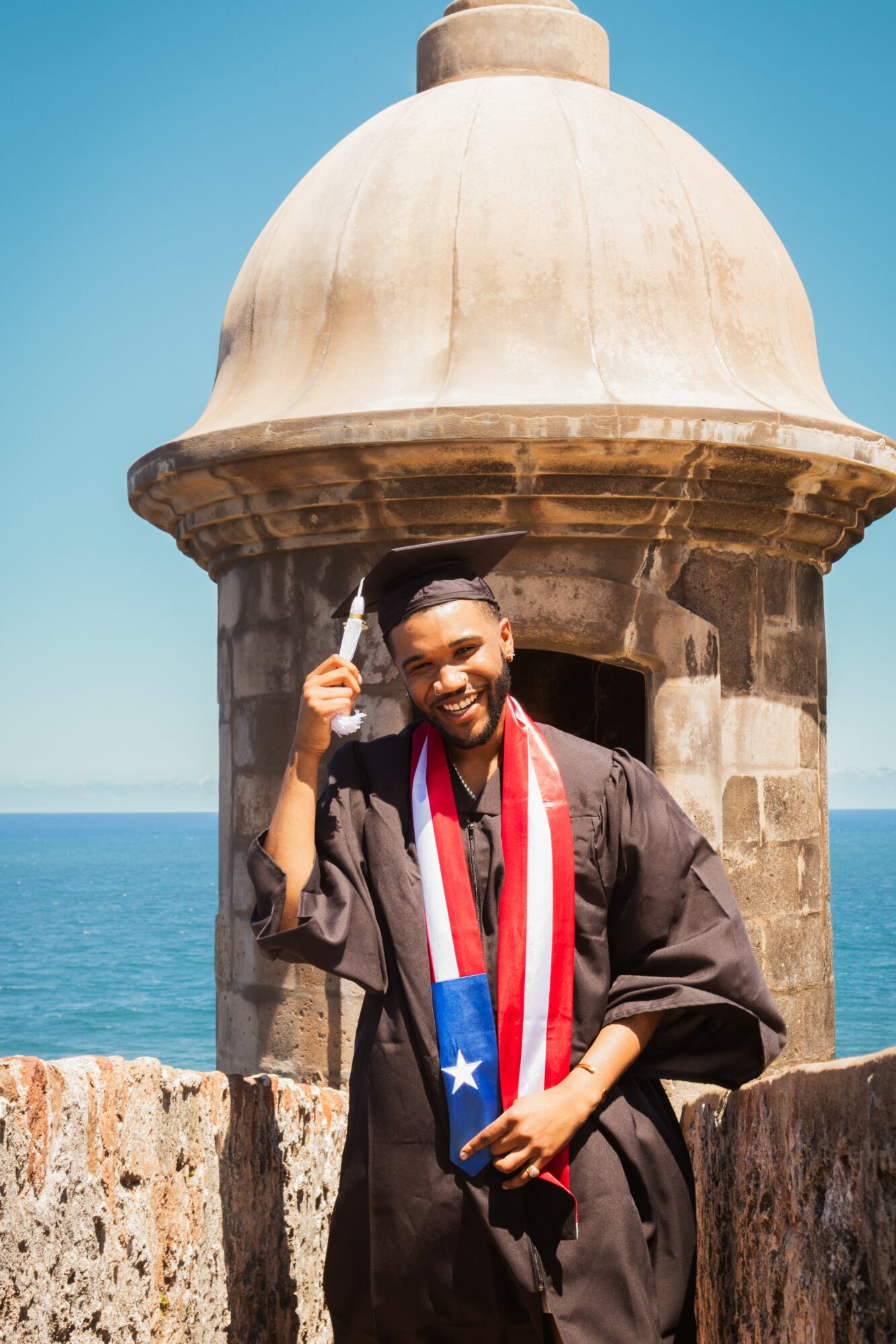 Graduate in cap and gown holding a diploma, smiling, with a lighthouse and ocean in the background.