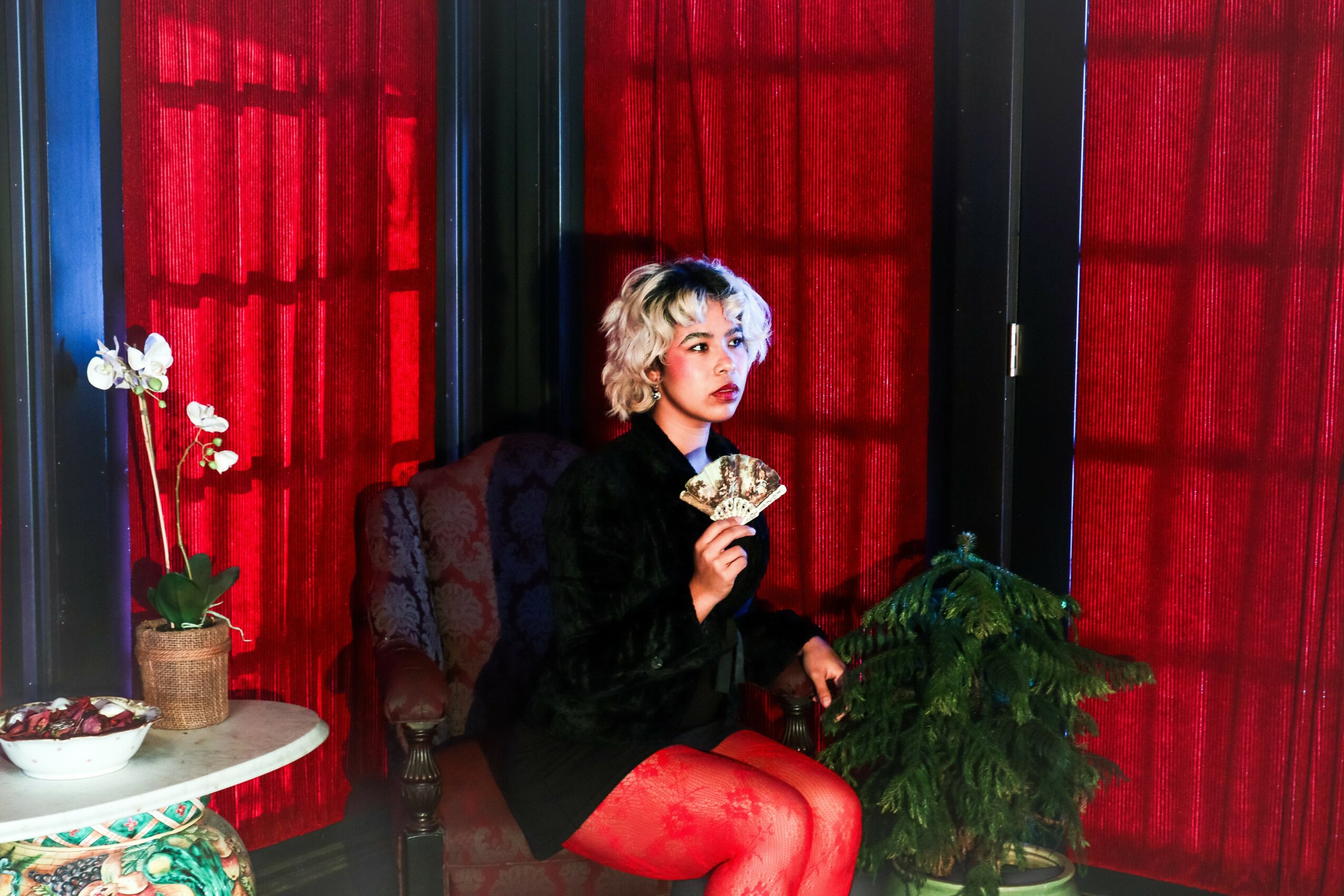 Woman with blonde hair and red lipstick sitting in a chair holding a fan, red curtains in background.