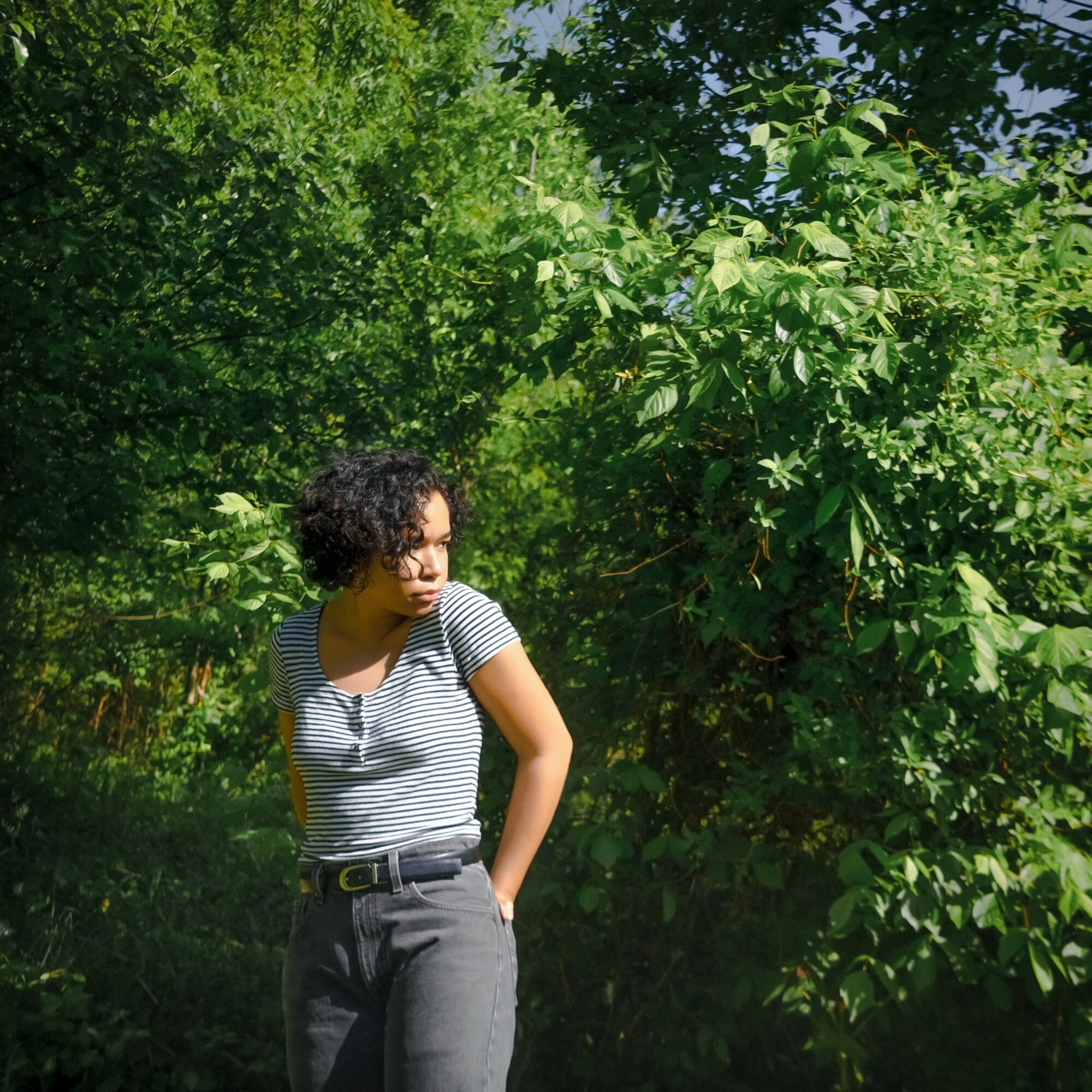 Young woman with curly hair in a striped shirt and gray pants stands outdoors among green trees and foliage.