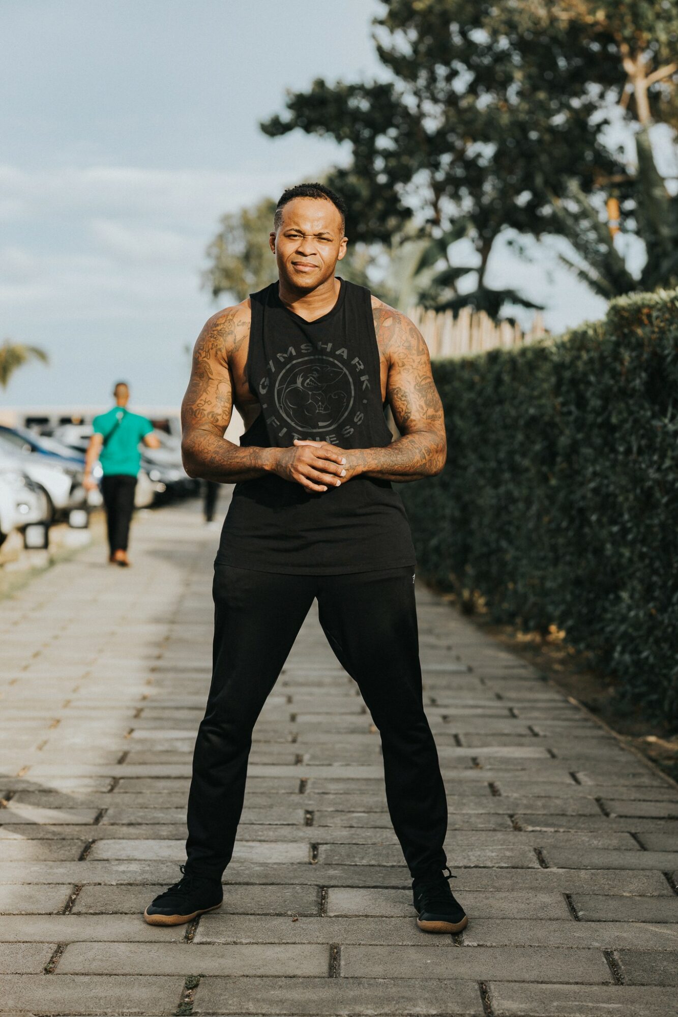 Man standing outdoors on a paved path, with trees and a person in the background, smiling and wearing a sleeveless shirt and black pants.