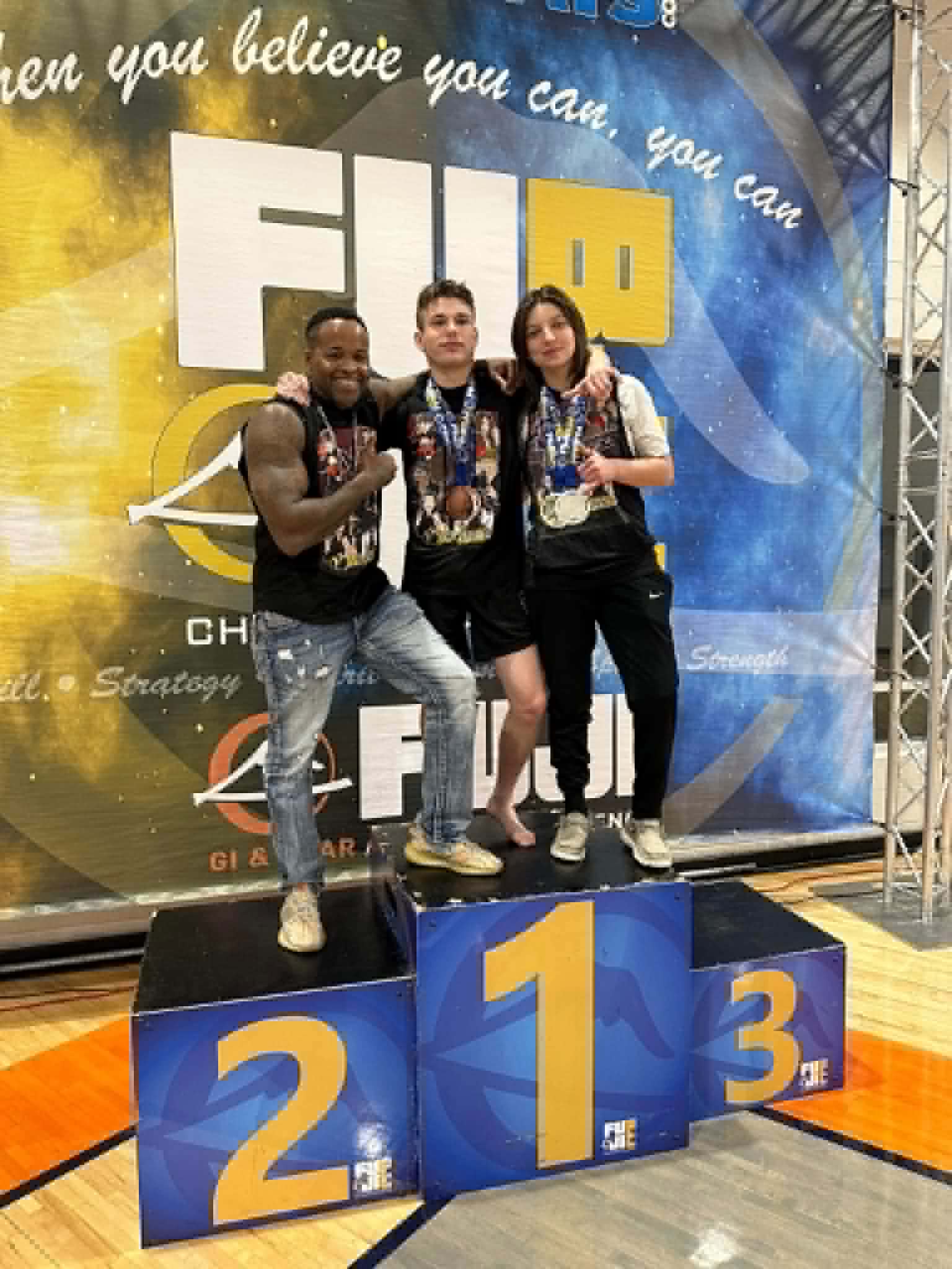 Three people standing on a winners' podium with numbers 2, 1, and 3, in front of a large FIRO banner. All are wearing medals.