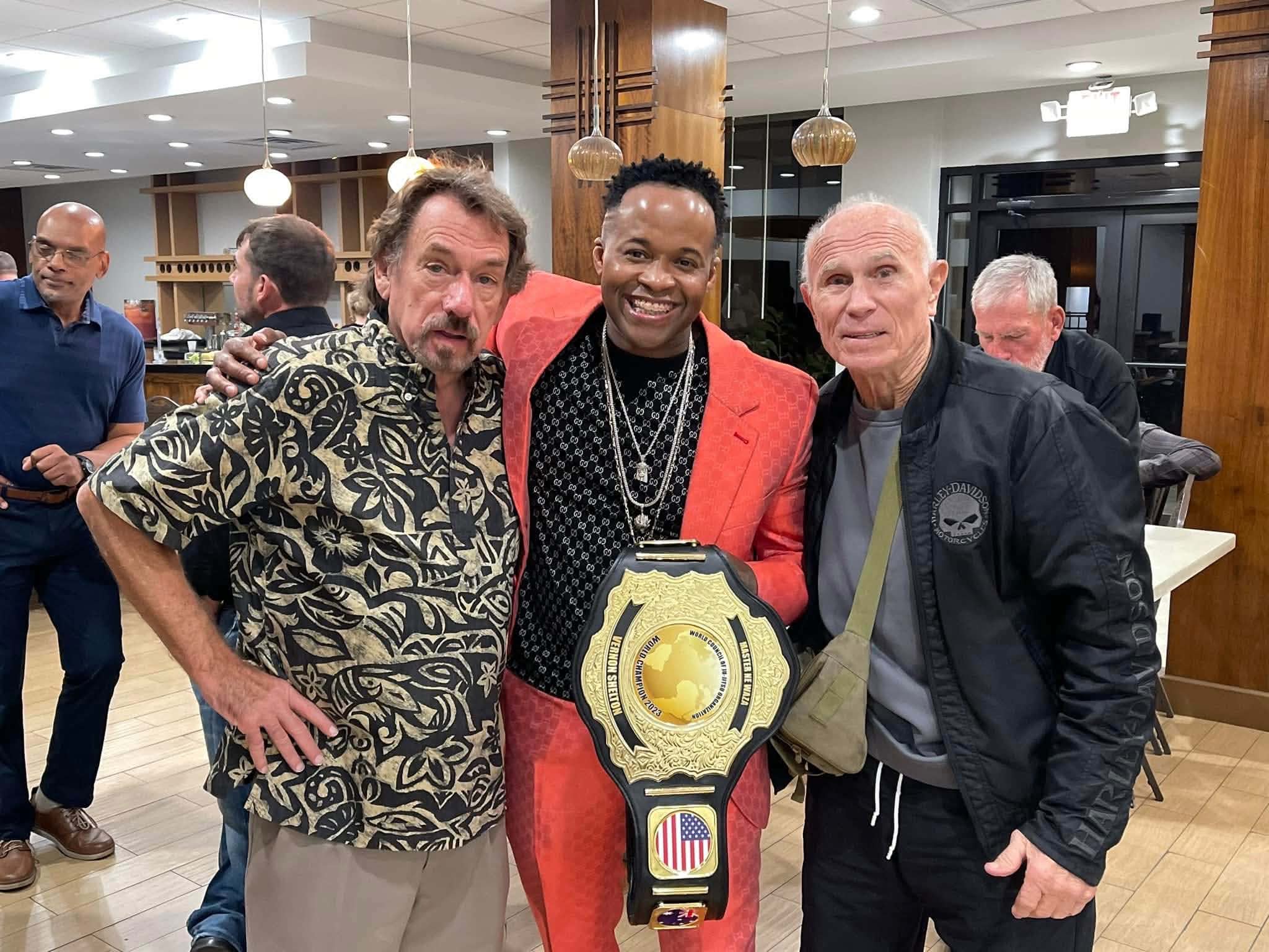 Three men standing together indoors, smiling, with a championship belt in front of them.