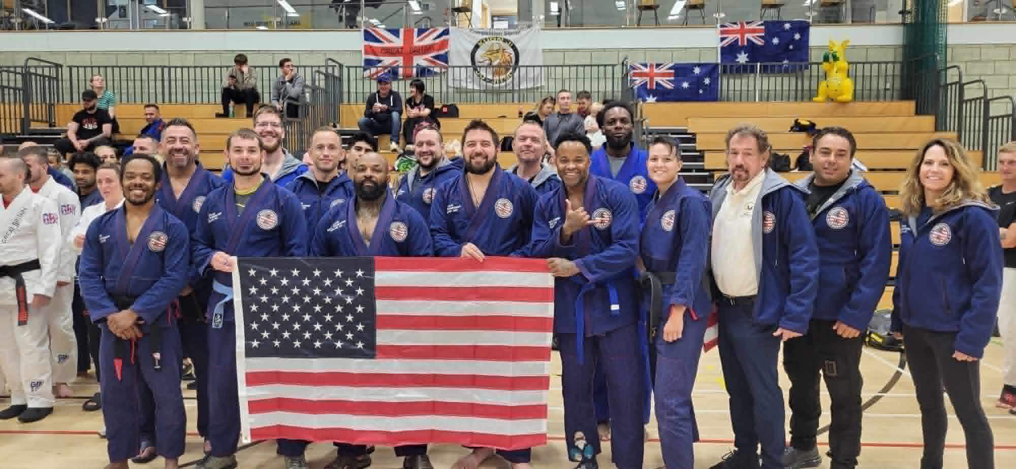 Group of people in martial arts uniforms holding an American flag, standing in a gymnasium with flags in the background.