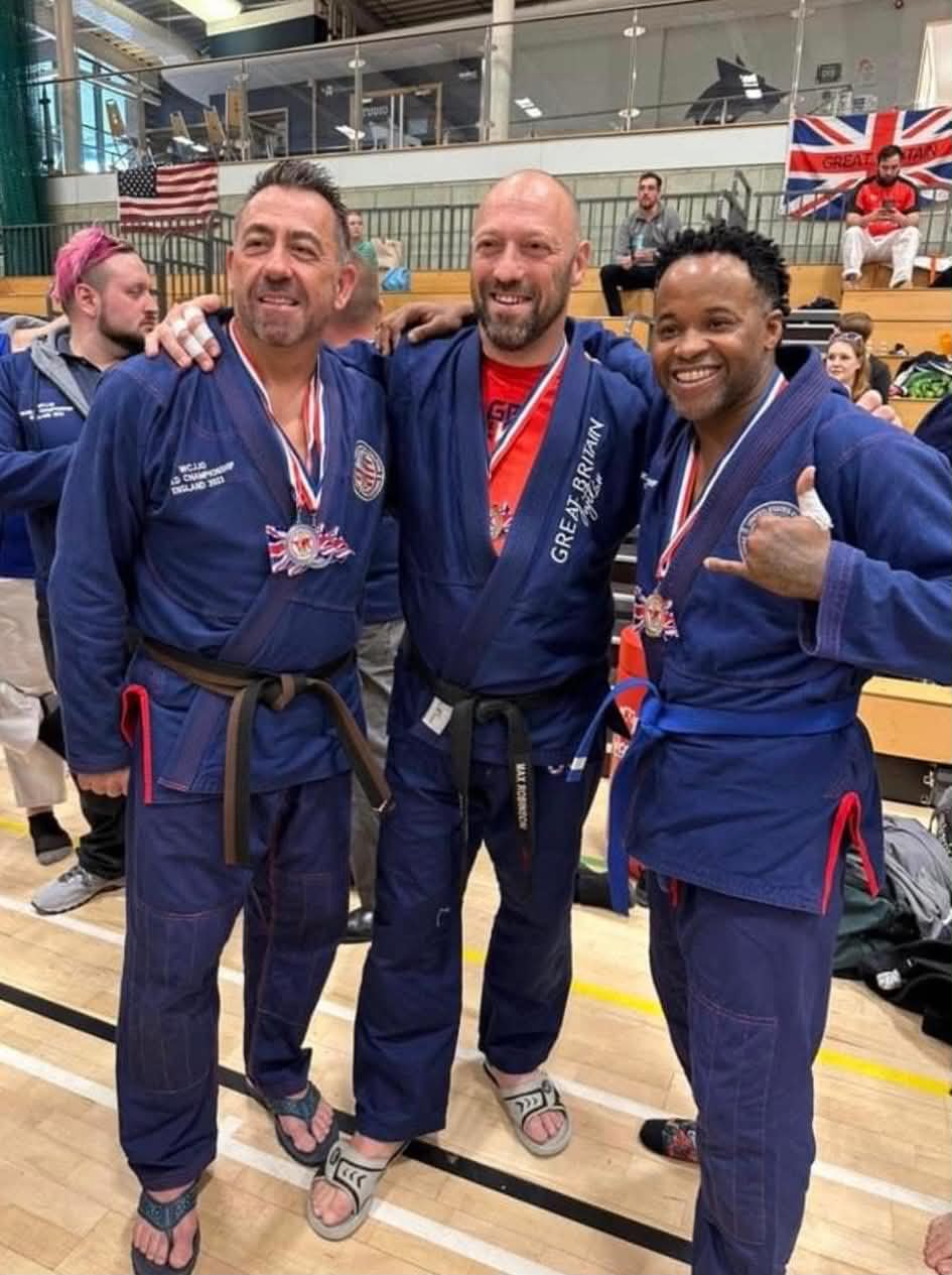 Three people in blue martial arts uniforms smiling and posing together in a gymnasium, with a British flag in the background.