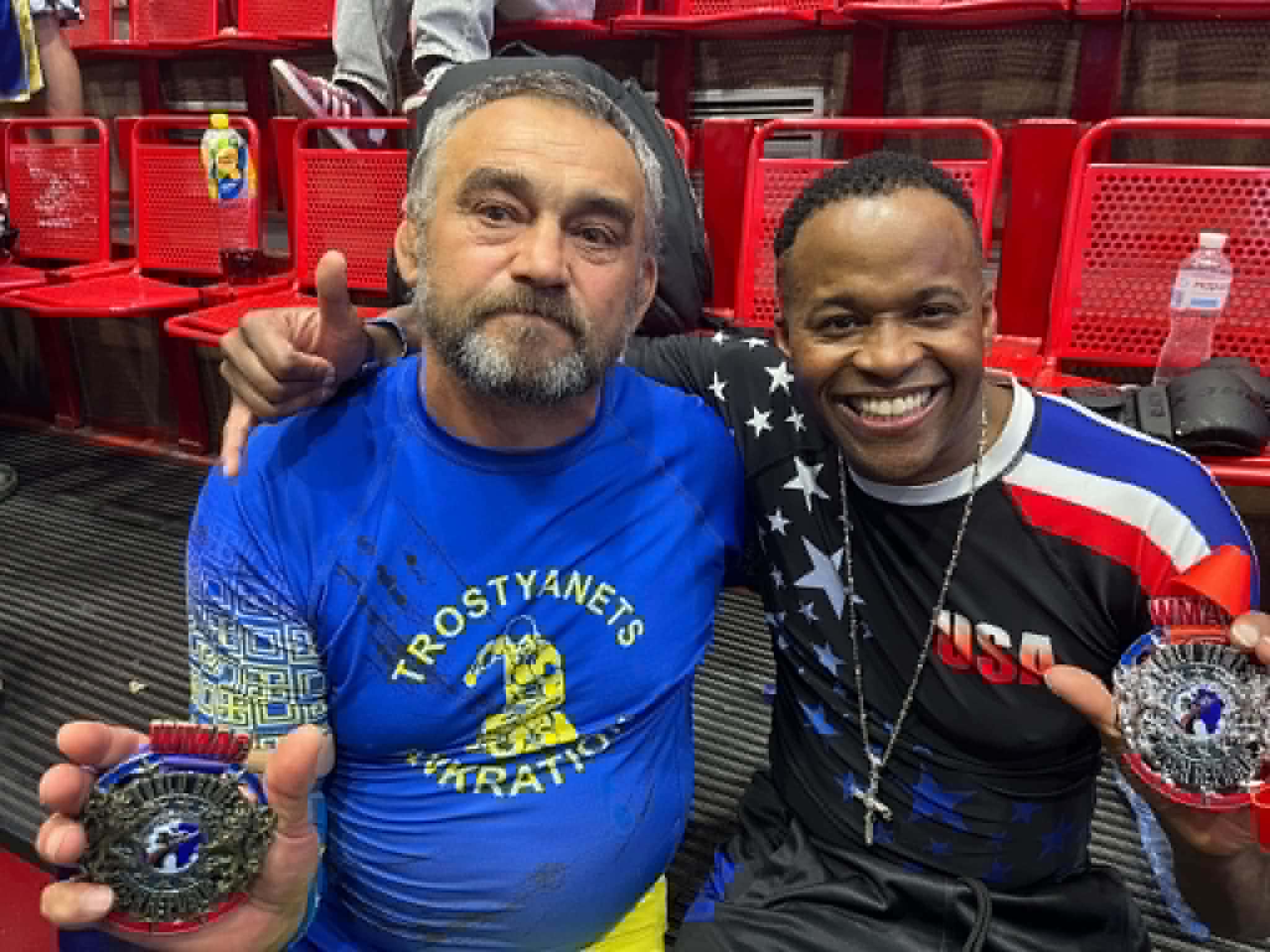 Two men smiling, holding medals, sitting in red chairs at an indoor event, one wearing a blue shirt, the other a black and red shirt.