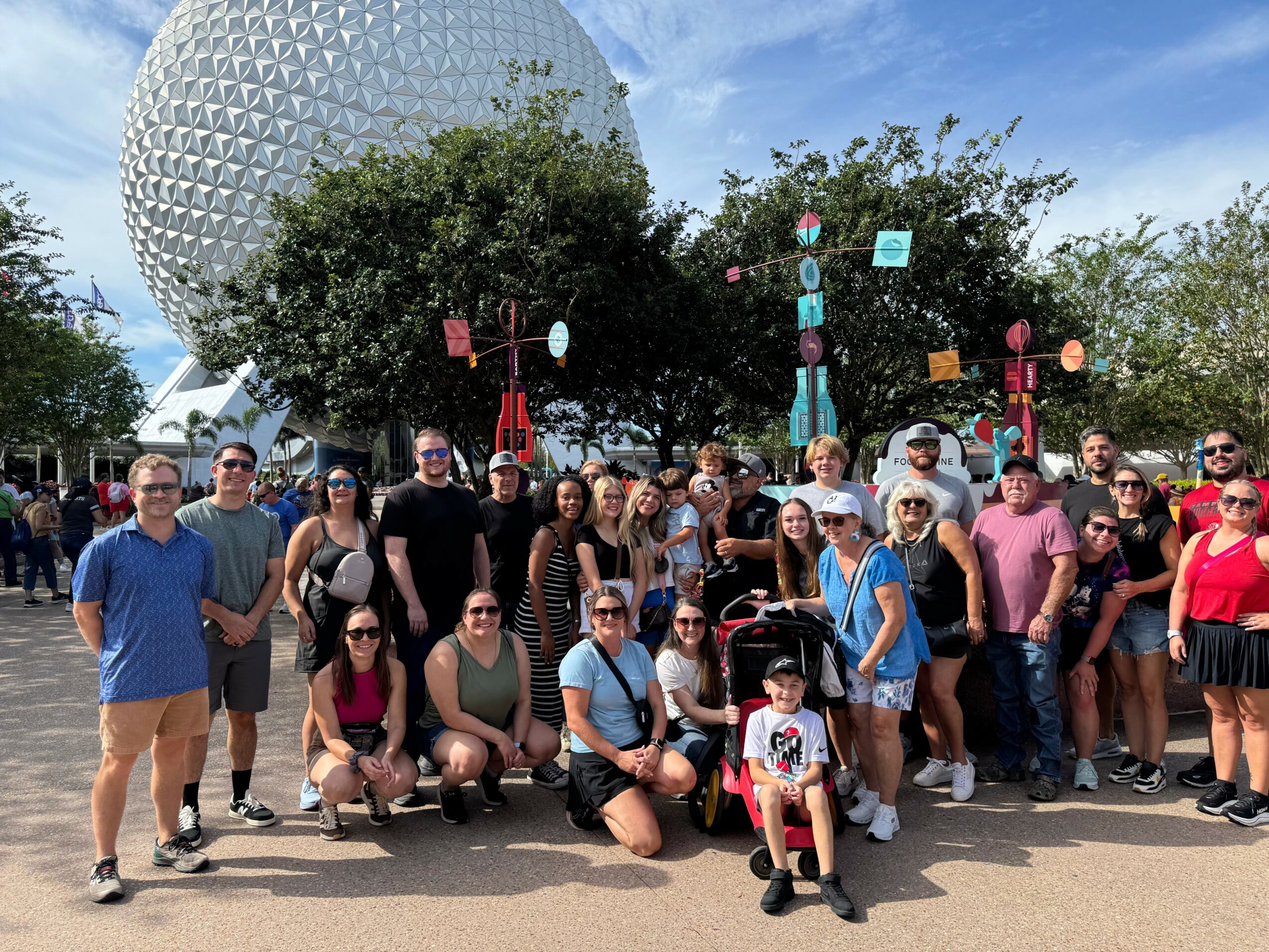 Group of people posing in front of a large geodesic sphere at a theme park, with trees and blue sky in background.