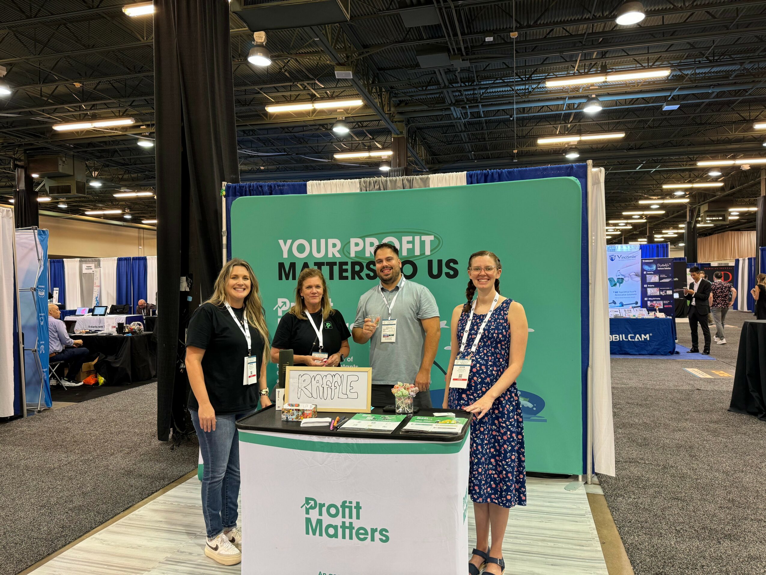 Four people stand behind a booth with a green and white sign that reads 'Profit Matters.'