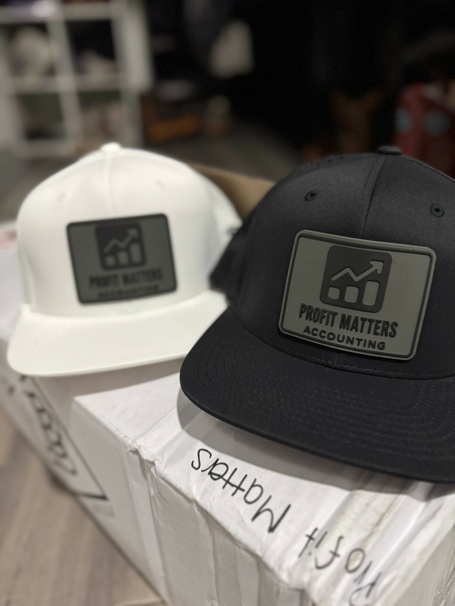 Two baseball caps, one white and one black, with a logo reading 'Profit Matters' on a box.