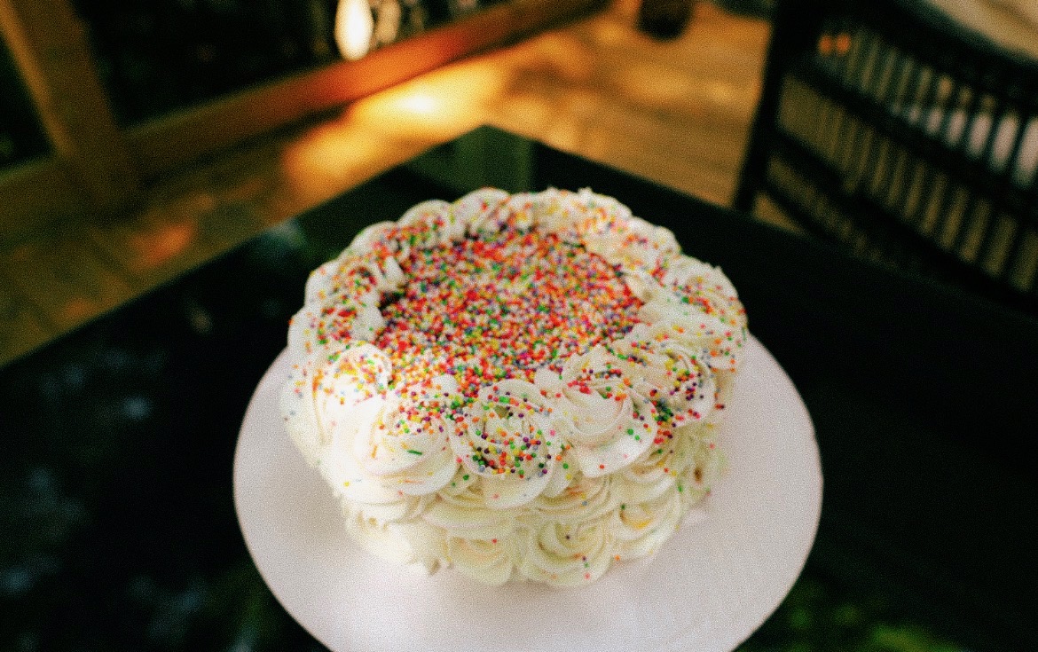 Round cake with white frosting, colorful sprinkles on top, decorated with piped cream, on a white plate.