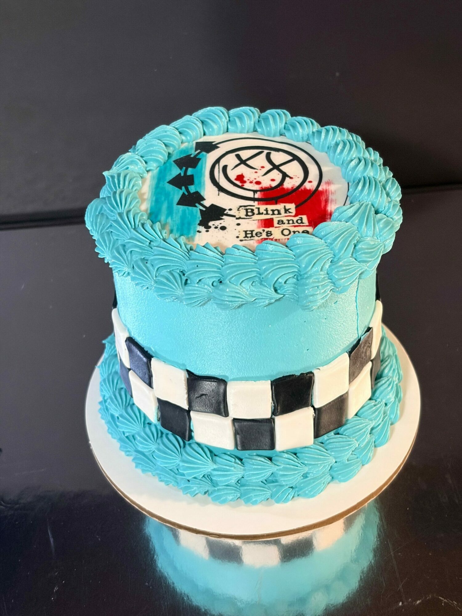 Two-tiered birthday cake with blue frosting, checkered pattern, and a birthday message on top.