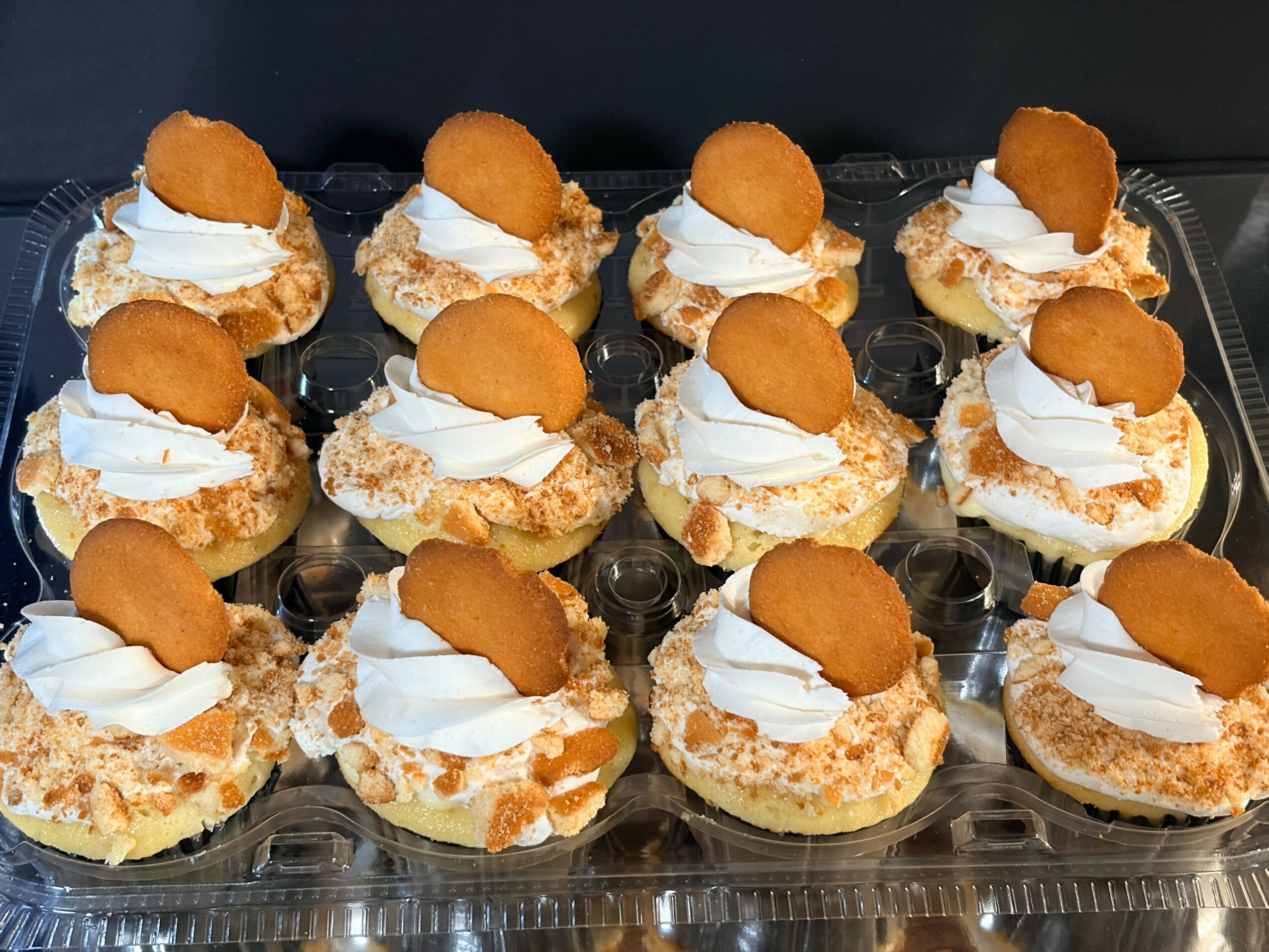 Twelve cream-filled pastries topped with whipped cream and a small round cookie, arranged on a clear plastic tray.