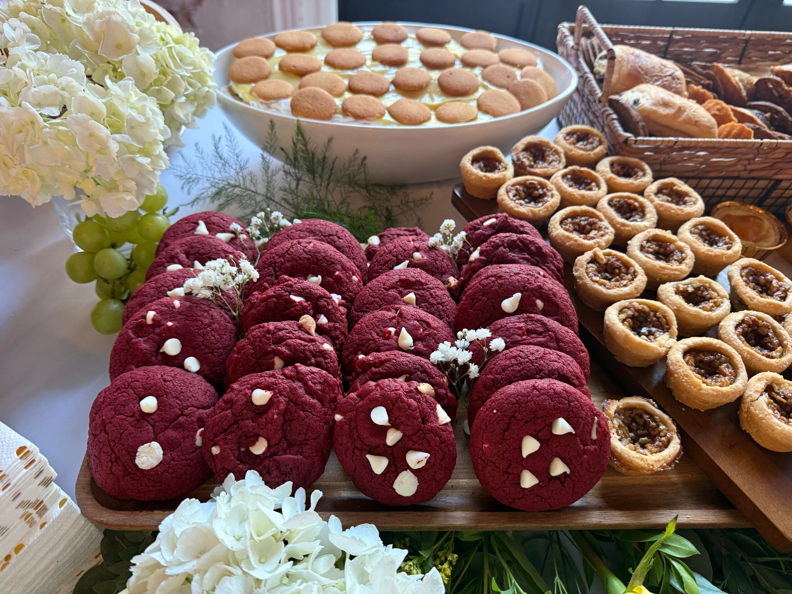 Assorted cookies and desserts on a table, including red velvet cookies with white sprinkles, small tartlets, and a bowl of macarons, with flowers and grapes nearby.