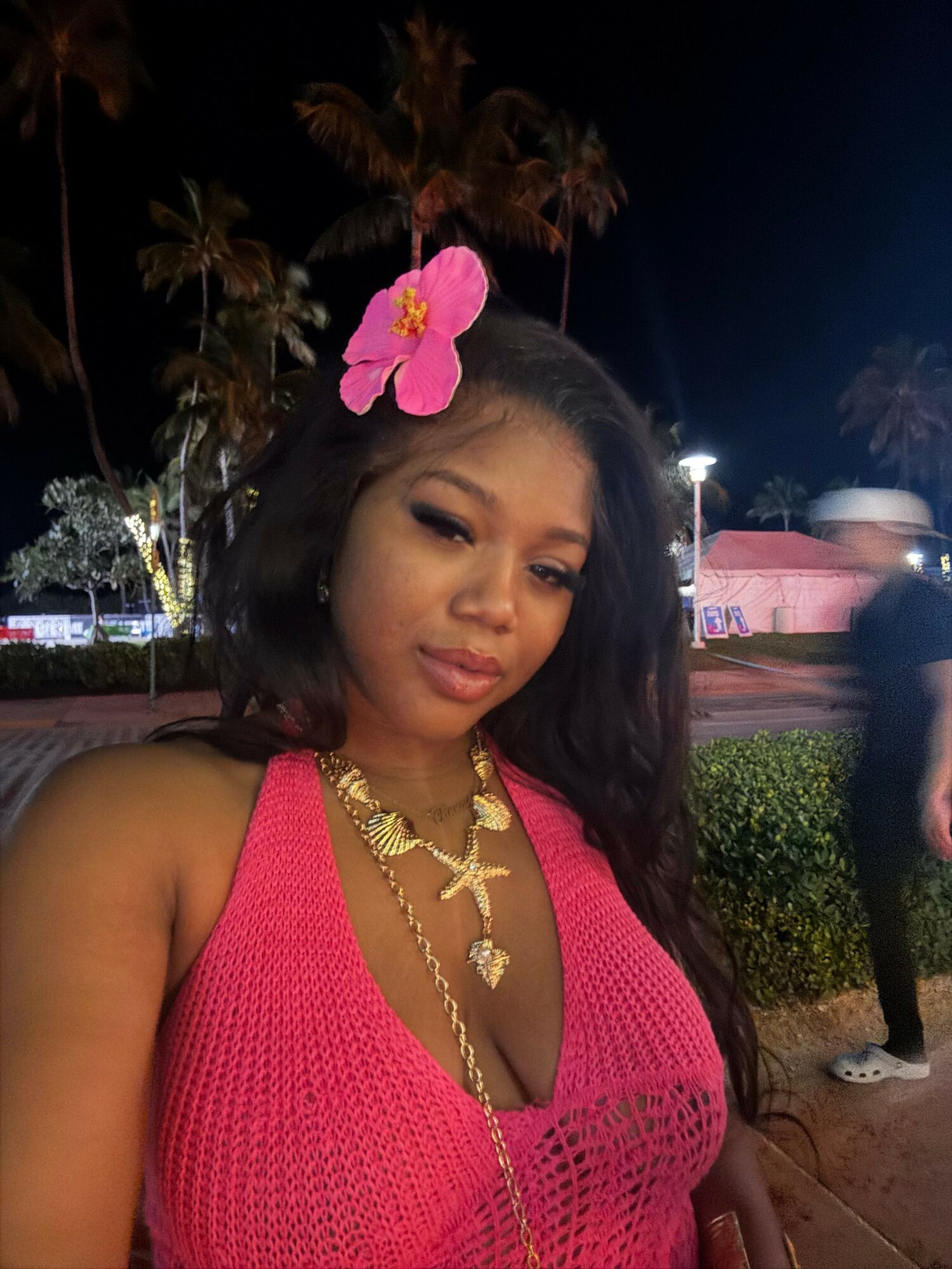 Woman with long dark hair wearing a pink top and gold jewelry, standing outdoors at night with a pink flower in her hair.