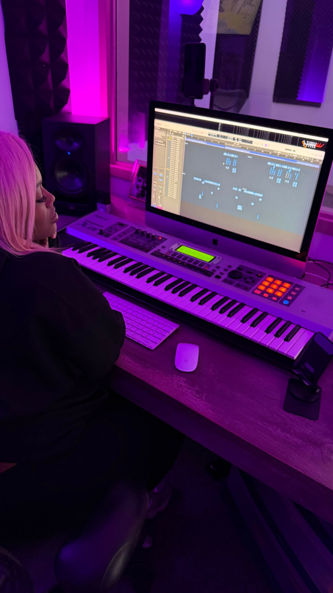 Person with blonde hair working on music production software in a purple-lit studio, surrounded by keyboards and equipment.