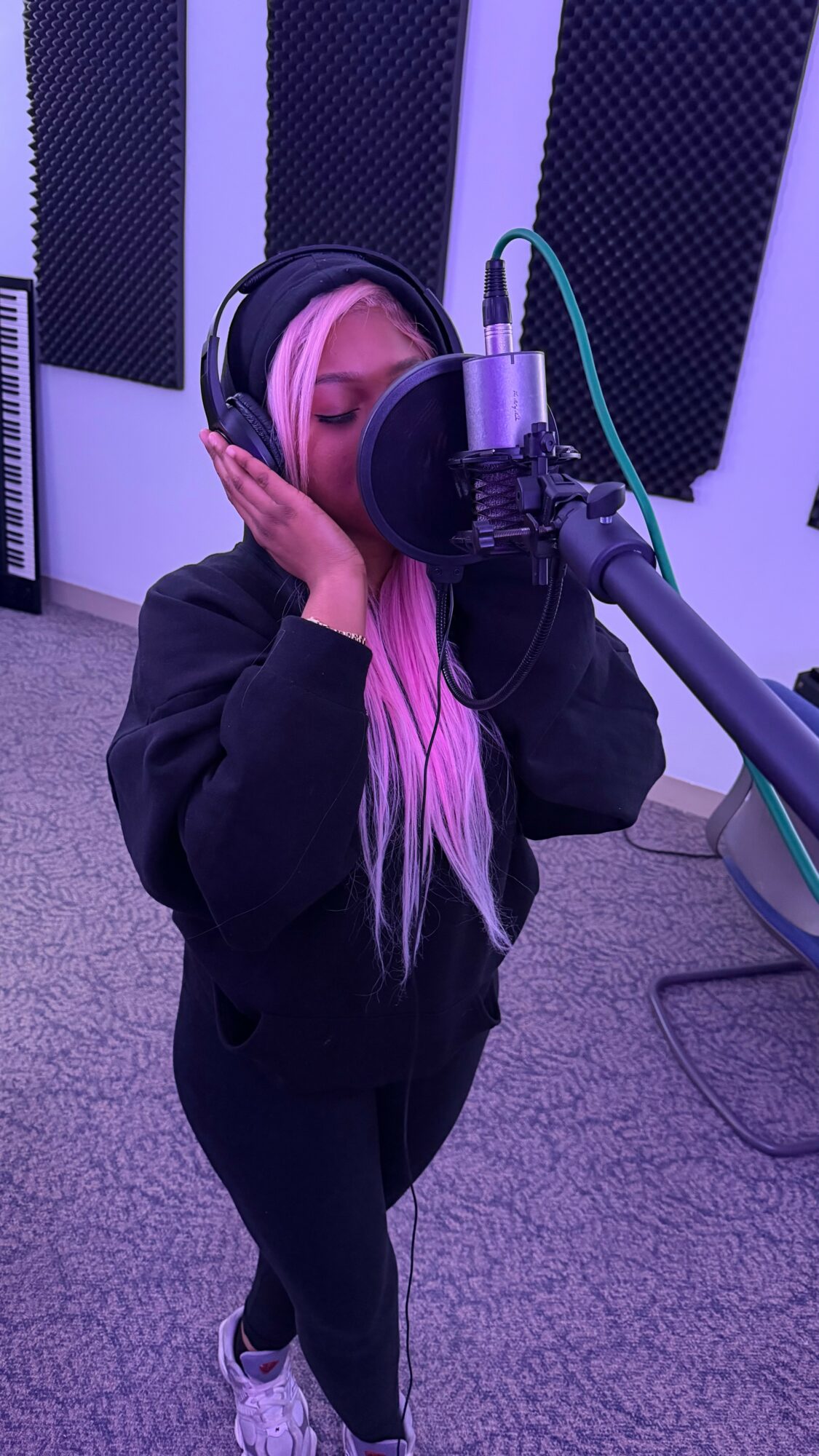 Person with long hair wearing headphones singing into a microphone in a recording studio.