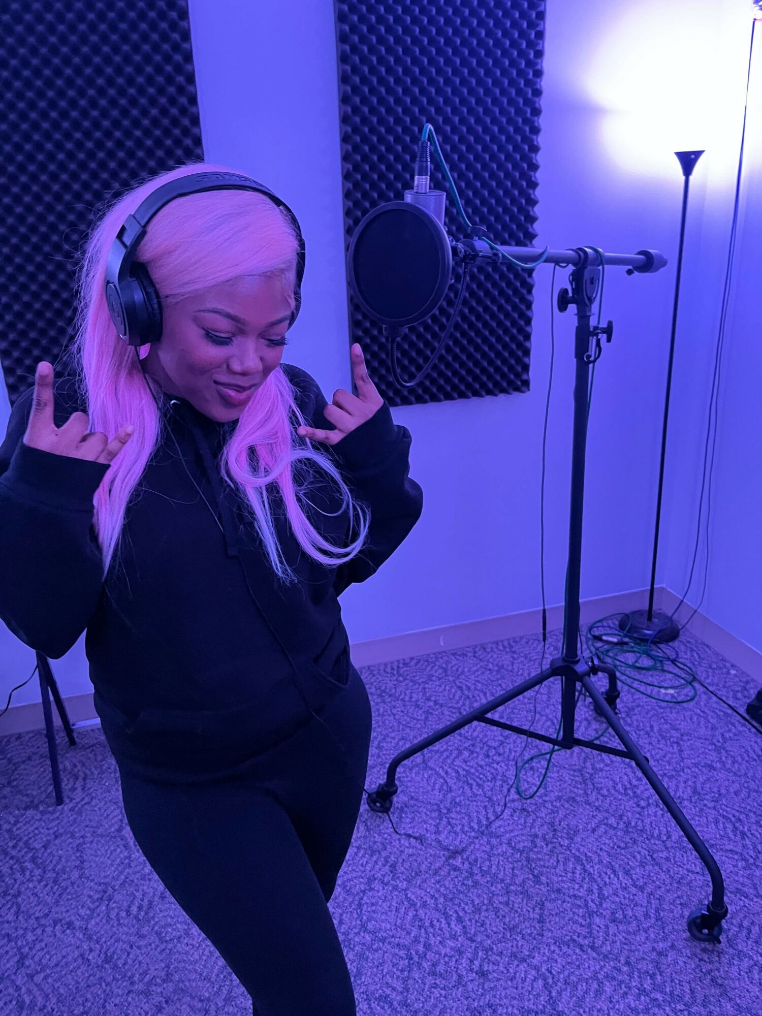 Woman with pink hair wearing headphones in a recording studio, standing with one hand raised, microphone and soundproofing behind her.