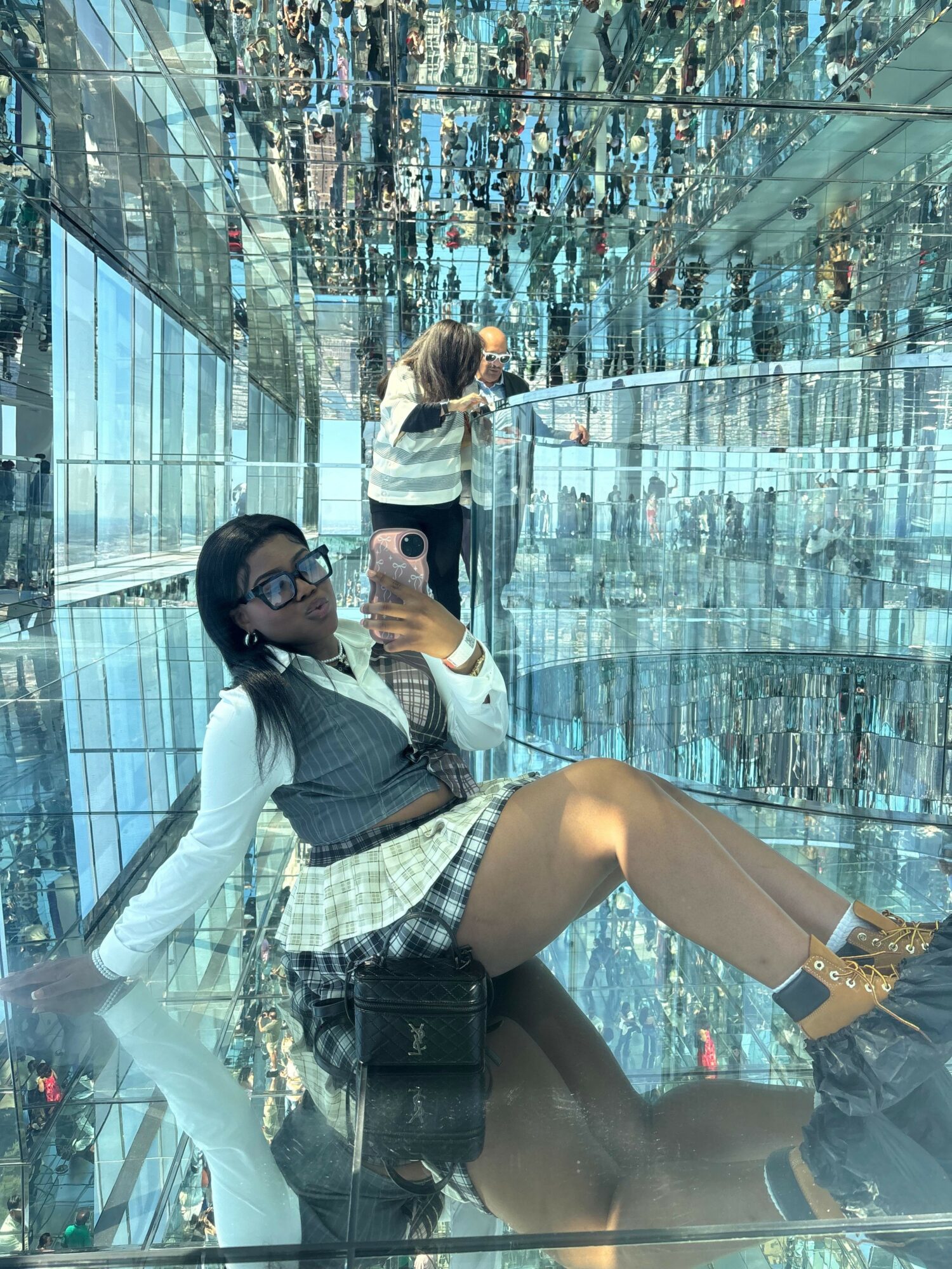 Person taking a selfie in a glass building with a city view, wearing sunglasses, a gray jacket, and shorts.