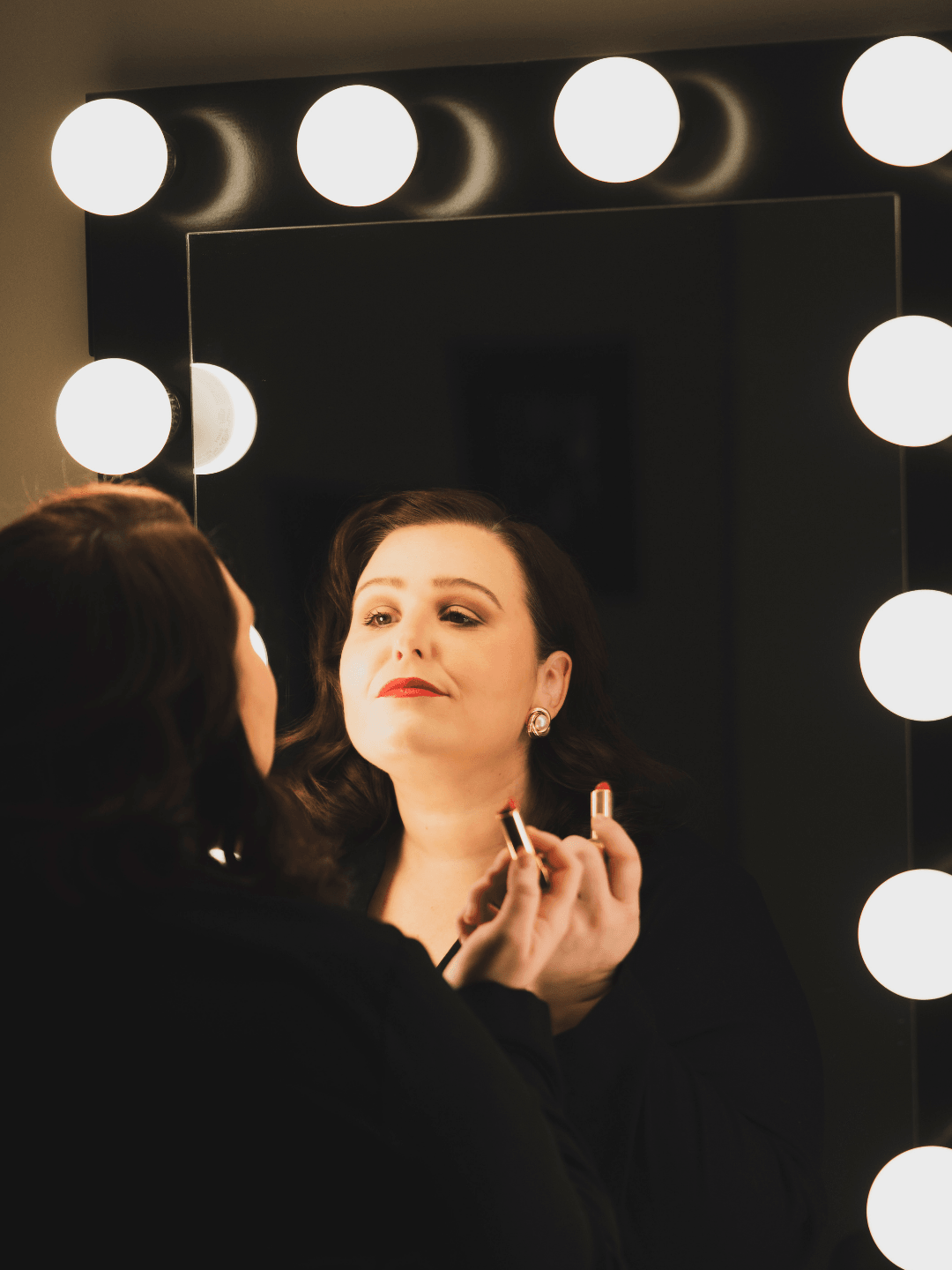 Woman applying lipstick in front of a mirror with bright round lights, reflection visible, dark background.