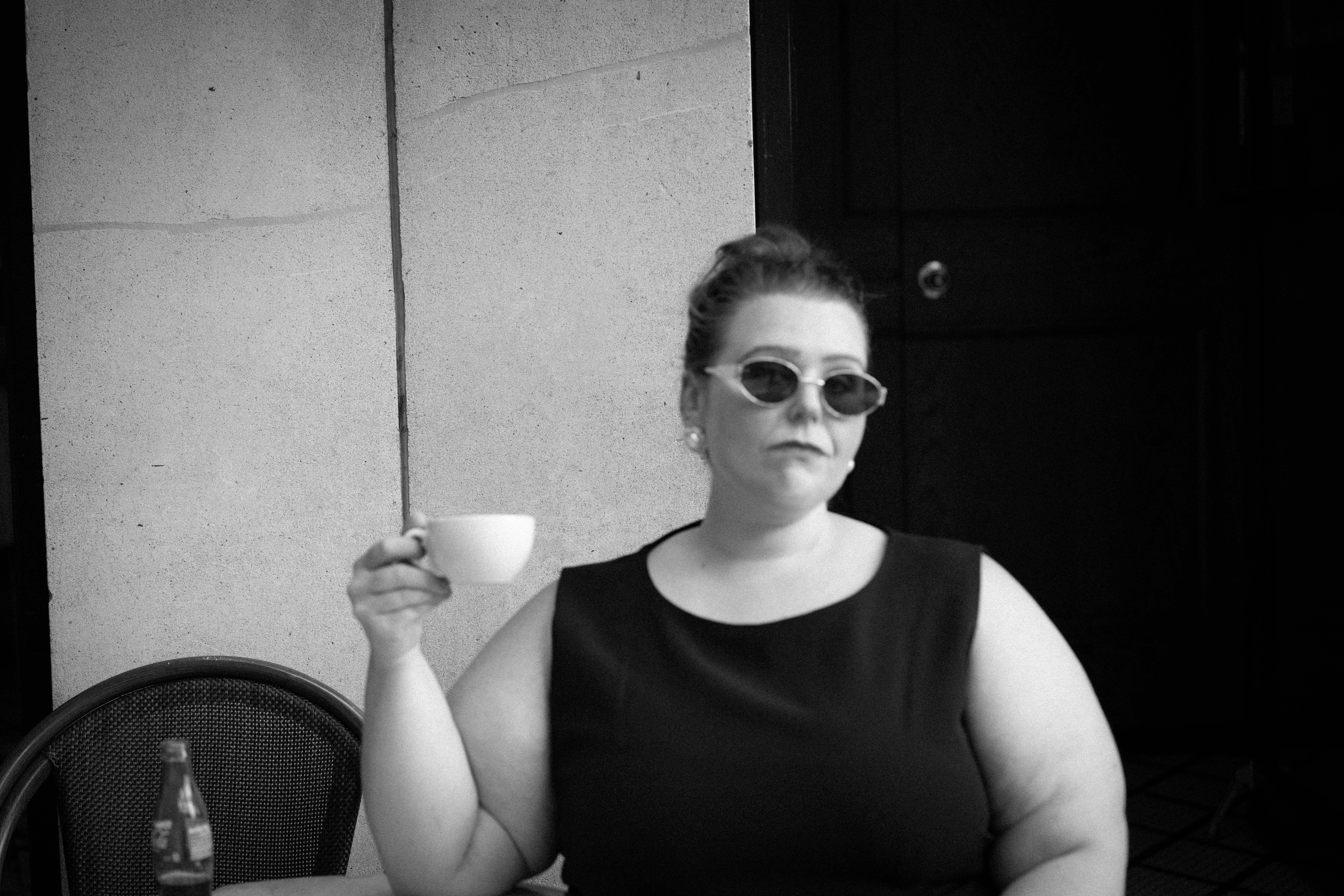 Woman wearing sunglasses and a sleeveless top, holding a small cup, sitting outdoors against a wall.
