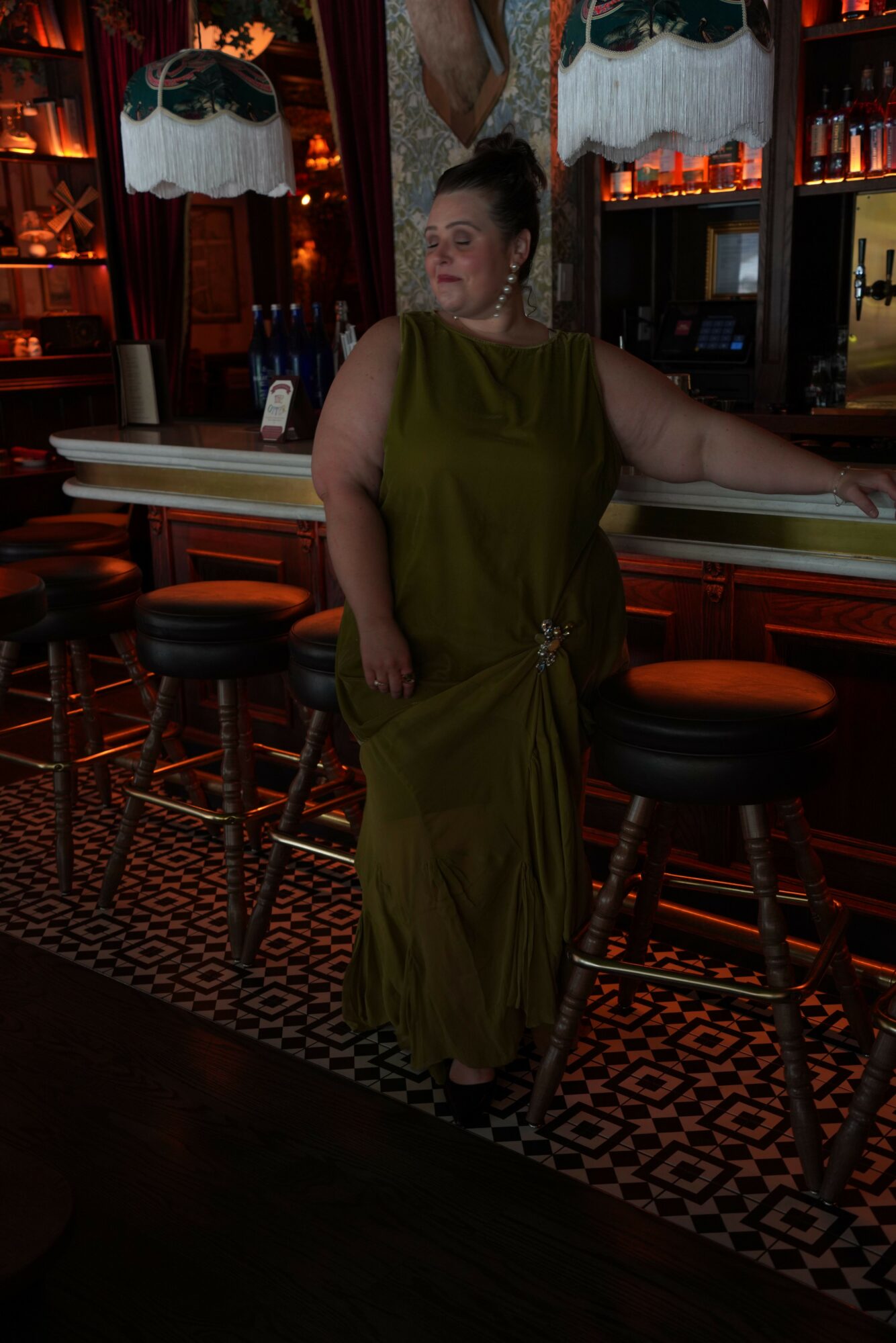 Woman in green dress standing at bar counter in a dimly lit bar with patterned floor and shelves of bottles.