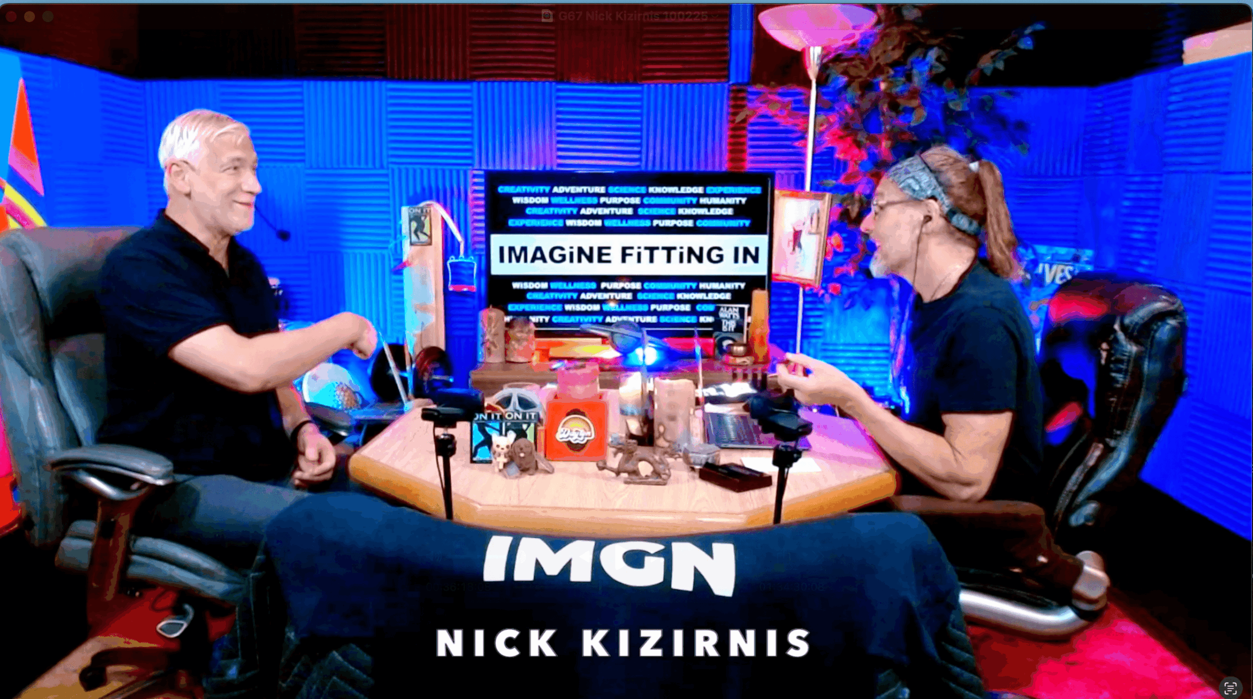 Two people sit at a table with equipment, smiling and talking in a colorful studio setting.