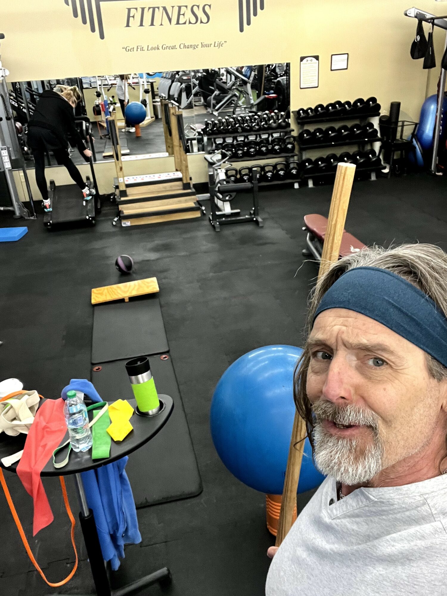 Man with headband taking selfie in gym with weights and exercise equipment in background.