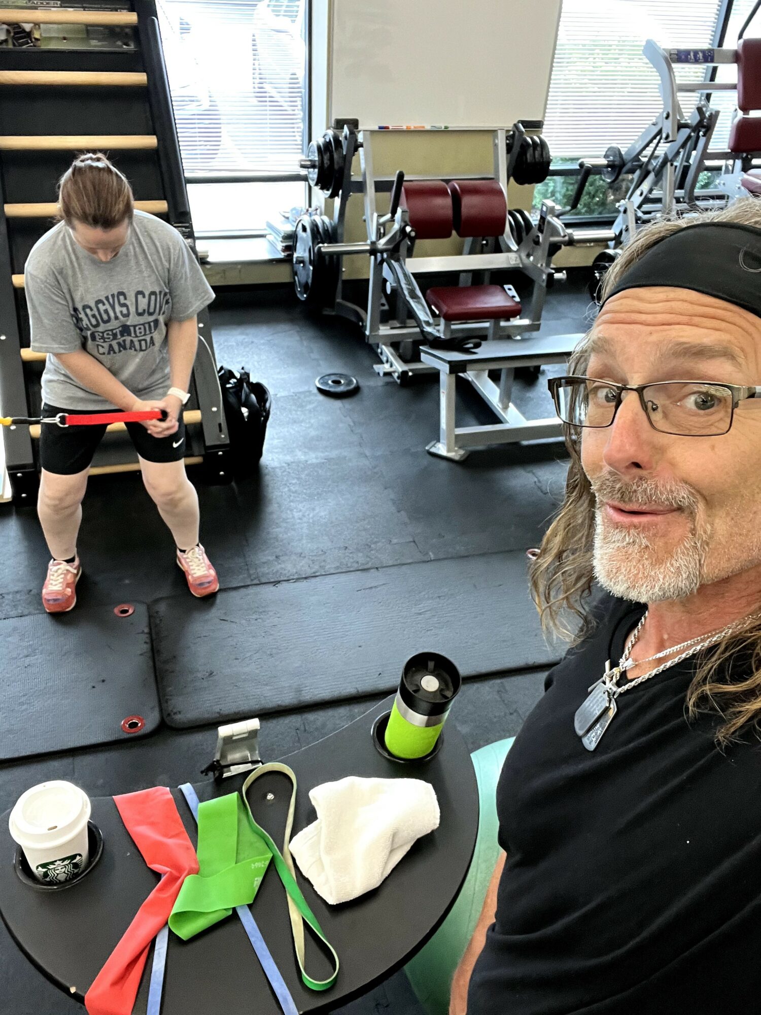 A man with glasses and a black cap takes a selfie in a gym, with a girl lifting weights in the background.